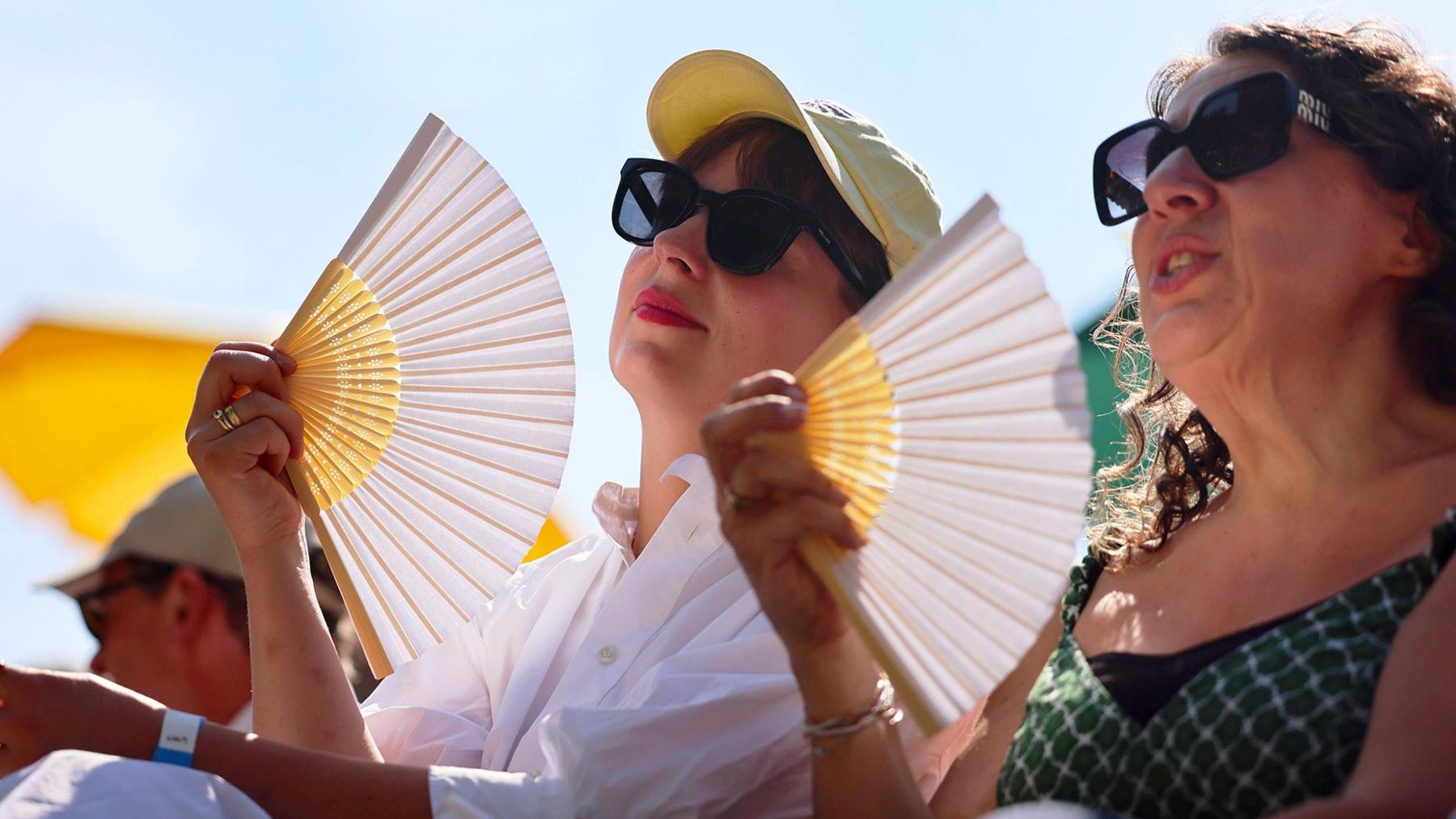 Two ladies in sunglasses sitting in the sun with matching yellow and white fans.