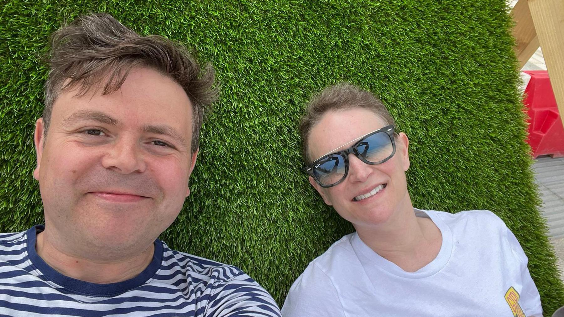 Geraint and Deb lying on grass. Geraint is wearing a navy and white stripy t-shirt and has short brown hair. Deb is wearing a white t-shirt and sunglasses and has her fair fair tied up.