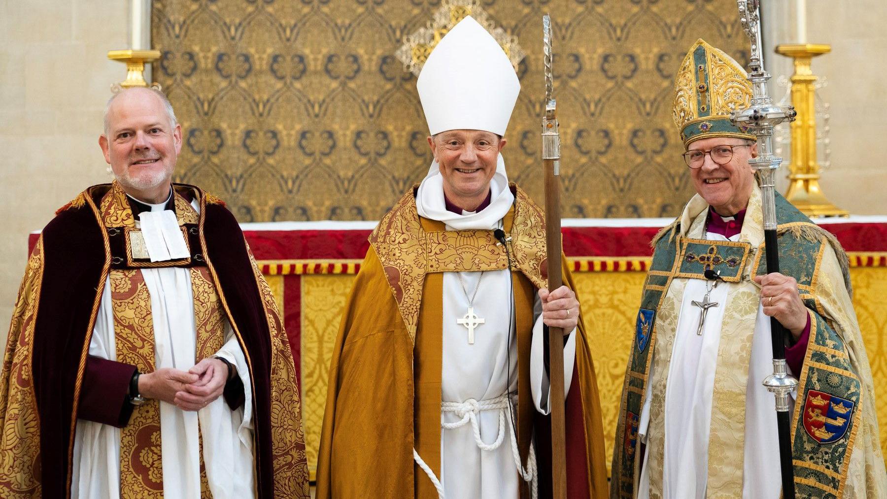 Joe Hawes, Mike Harrison and Martin Seeley stand in a row wearing their Anglican Pontifical vestments. They are all smiling at the camera and standing in front of an altar. 