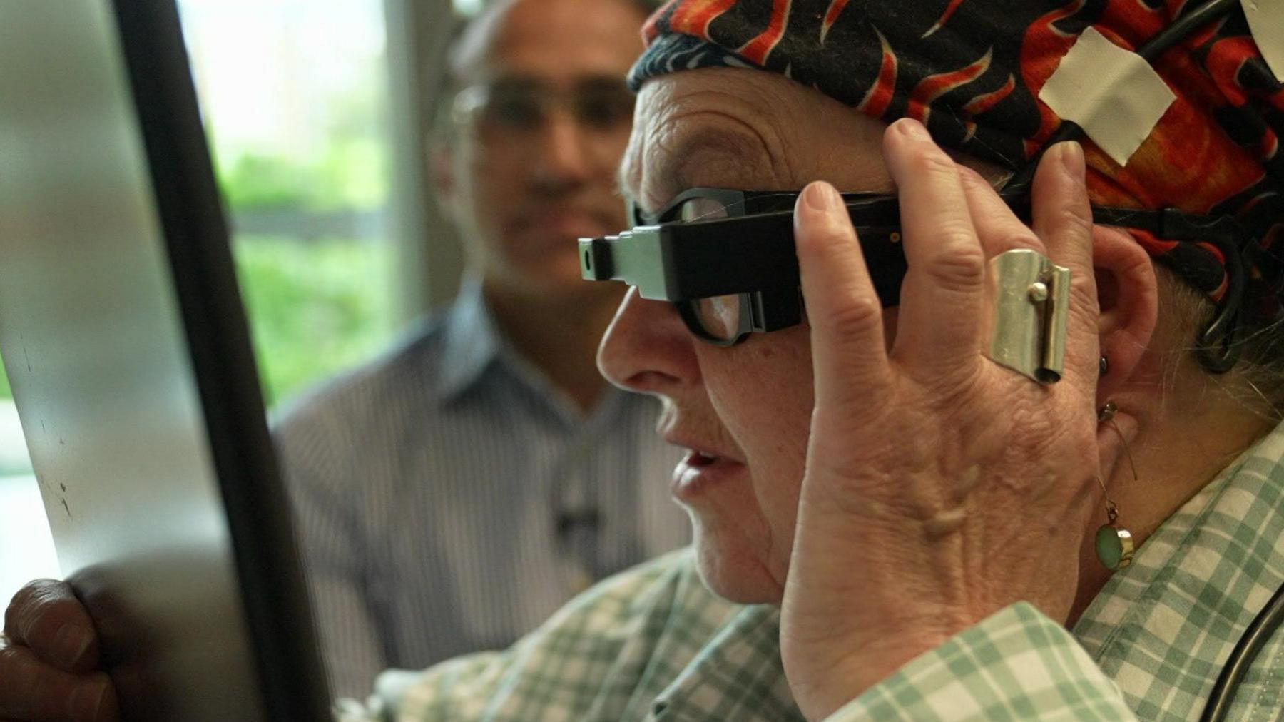 Sheila is wearing the special glasses, reading from a tablet a few centimetres from her face - she is side-on, holding one hand to the side of her face and concentrating hard. Watching behind her, slightly blurred, is consultant ophthalmic surgeon Mahi Muqit, from Moorfields Eye Hospital.