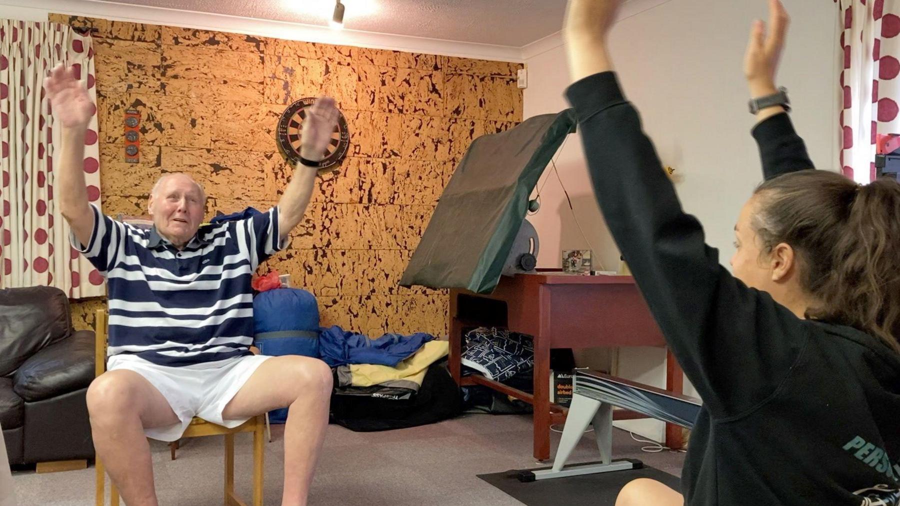 A man wearing a t-shirt with stripes and shorts on sits on a chair in a home gym. He is lifting his hands in the air. To the right, a woman wearing a black hoodie also puts her hands in the air. Other pieces of gym equipment can be seen in the background, as well as a darts board.