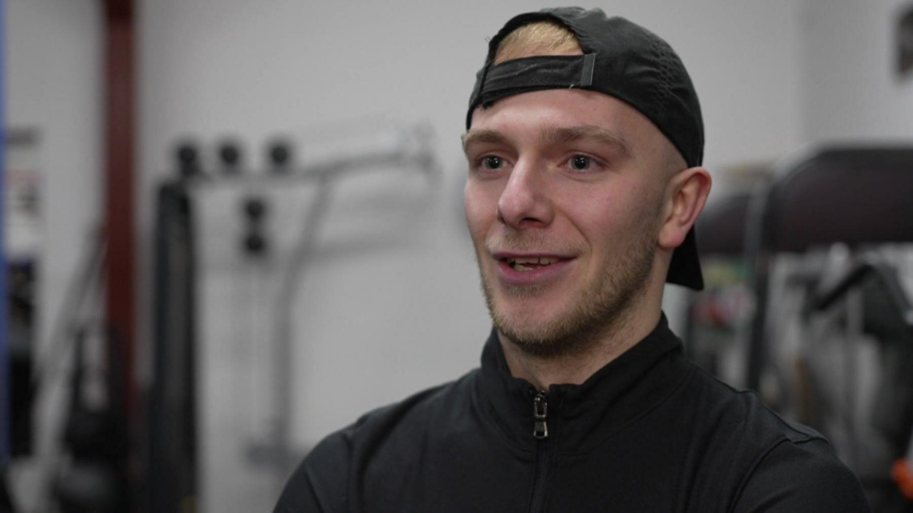 A man wearing a black cap backwards on his head and wearing a black zipped top is stood in a boxing gym talking to the camera.