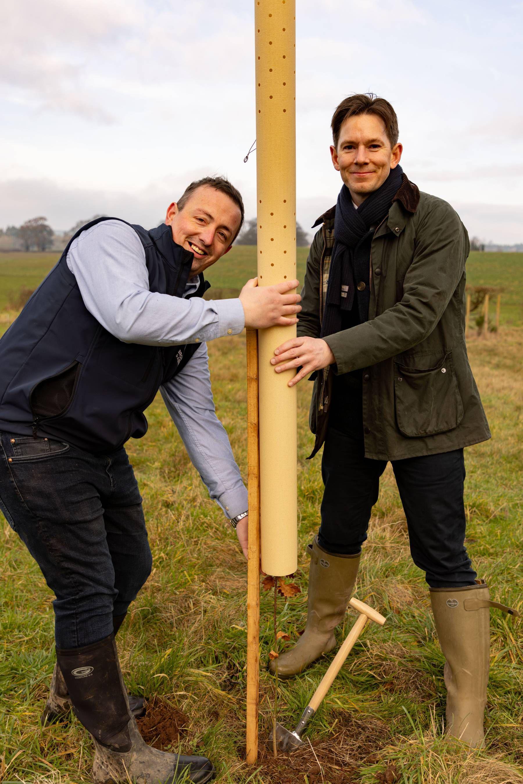 Two white men, one wearing dark trousers, wellies, a light blue shirt and a blue body warmer holding a yellow tree guard while the other man holds the other side wearing green wellies, dark trousers, a green jacket and black scarf. they are standing in a field 