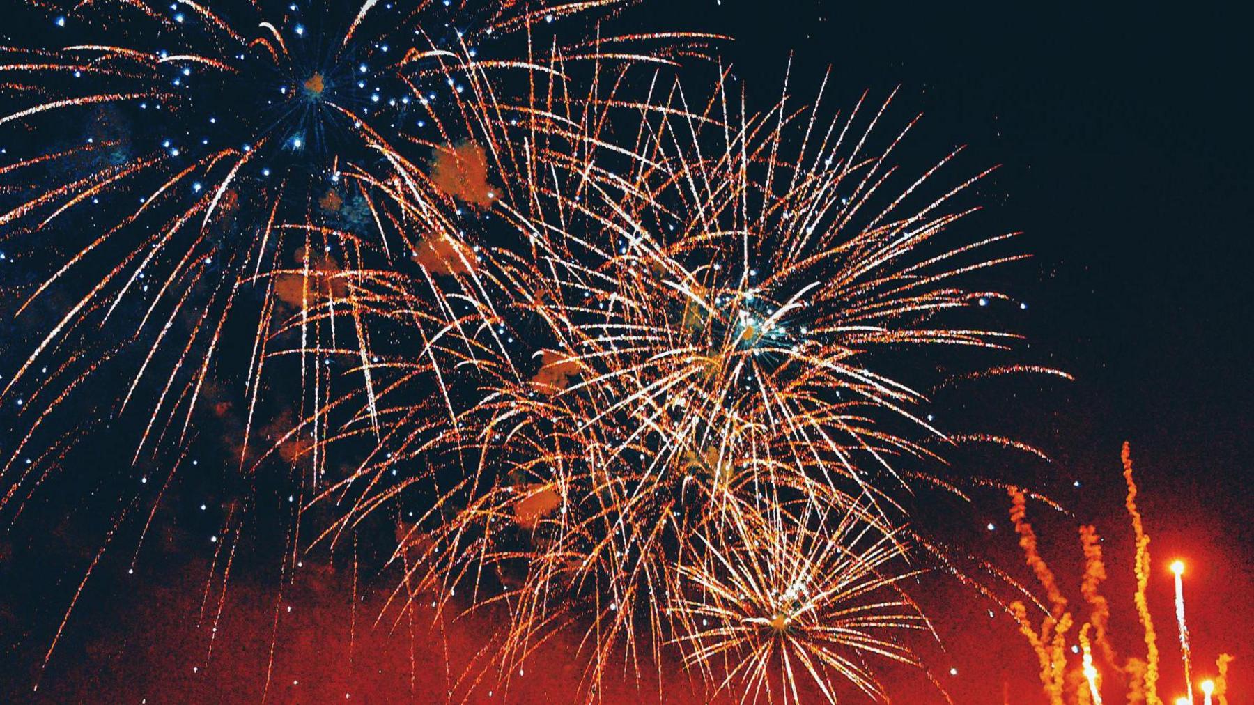 Starbursts of orange and yellow fireworks against a dark sky, with red smoke and lights rising to the right.