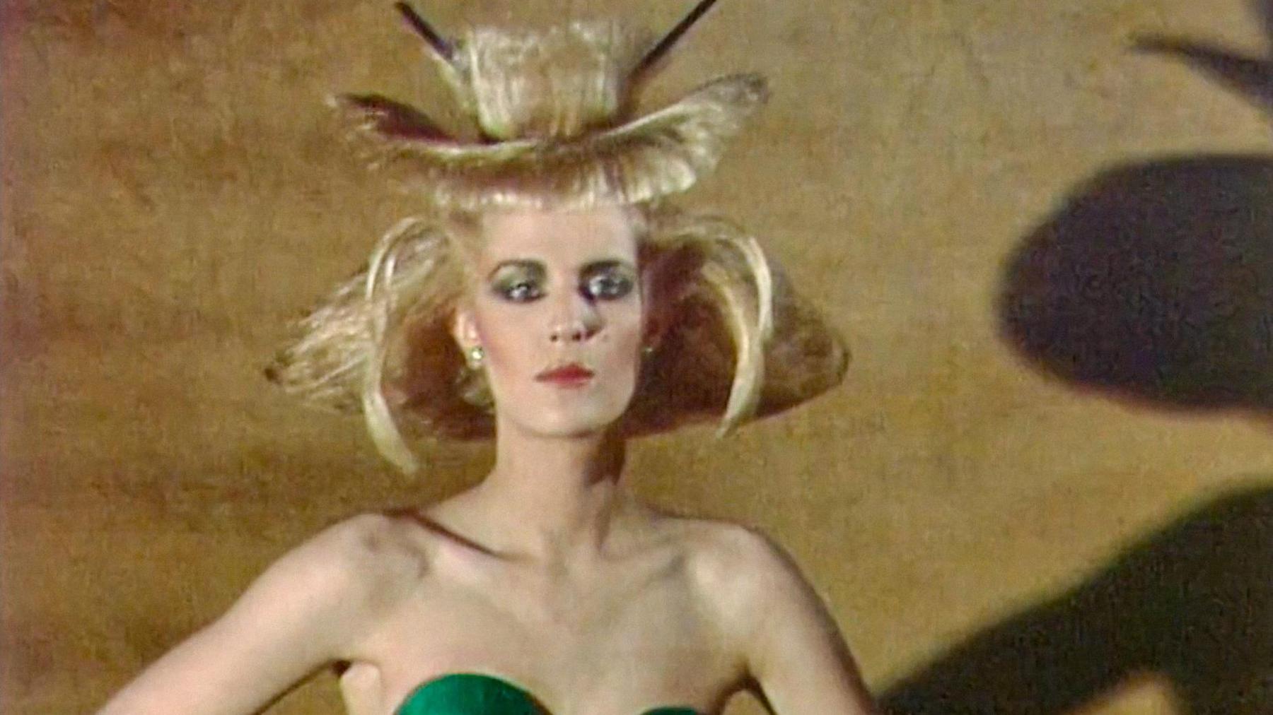 A woman in a green bustier and hair shaped like an orchid stands in front of a brown wall