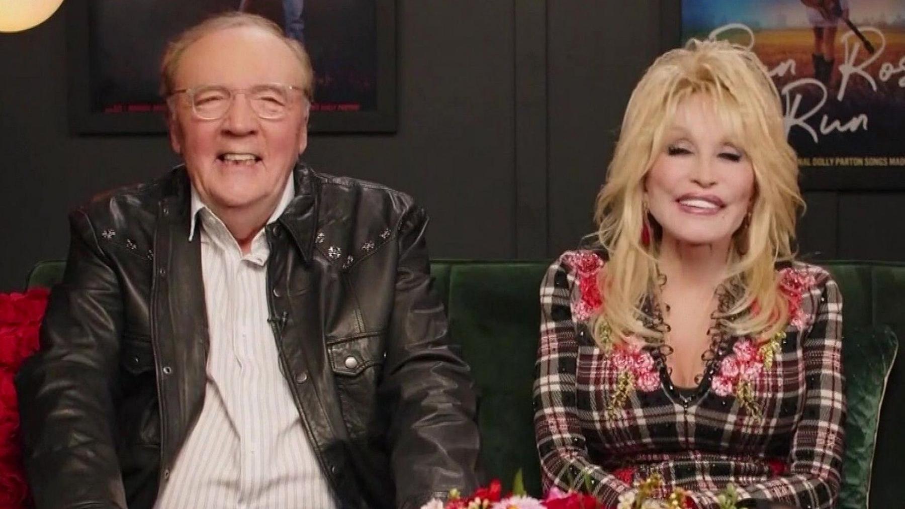 James Patterson, seated to the left, has thin, receded greyish hair, and wears glasses, a shirt and leather jacket, and is smiling. Dolly Patron has blonde curled hair and is wearing a low-cut checkered dress and is smiling. 