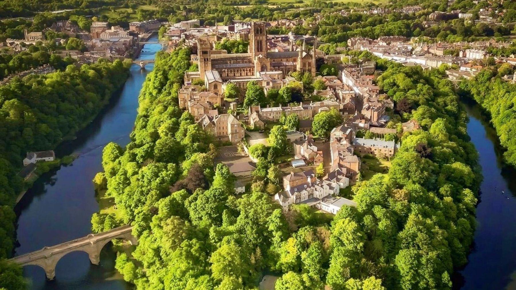 An aerial view of Durham City Centre. The grand cathedral and surrounding stone buildings sit on a peninsula that is bordered by lush green trees.