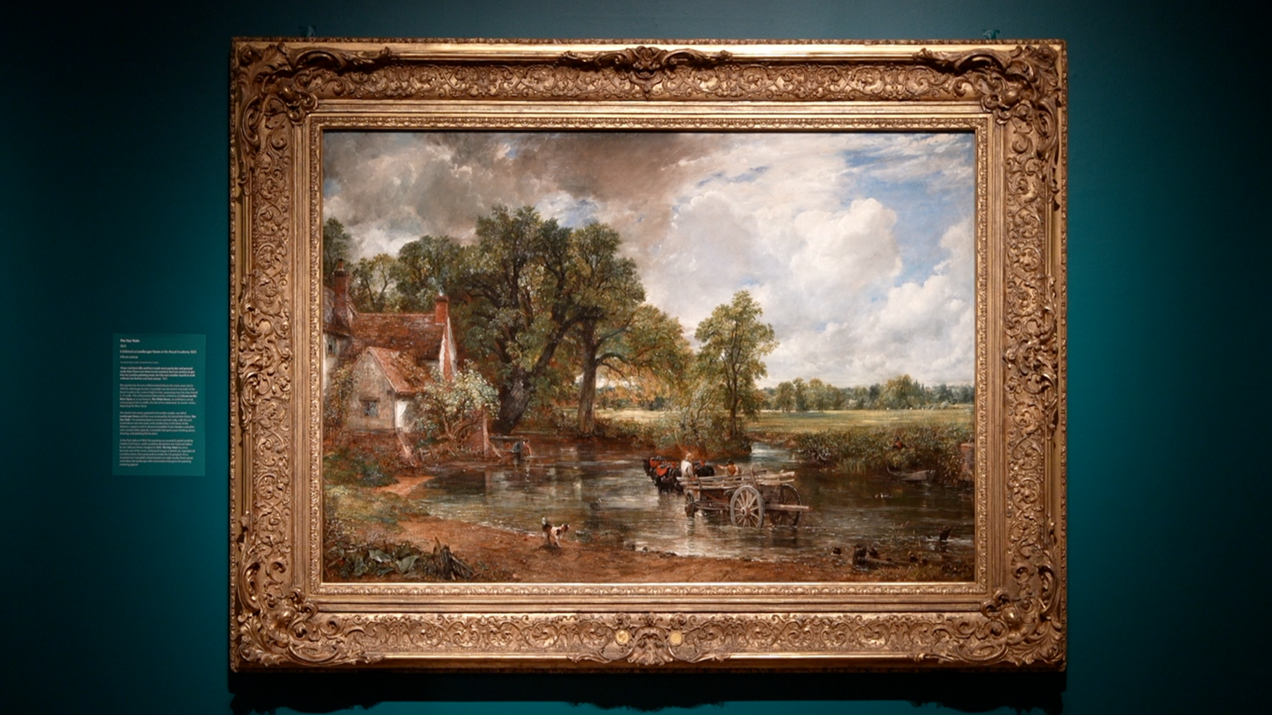 A large oil painting displayed on a wall as part of an art exhibition. The painting depicts a rural scene with a horse and cart moving through a river. There is a cottage on the bank of the river. 