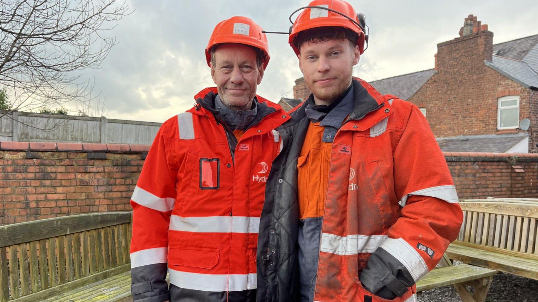 Two men dressed in orange hi-vis clothing with a logo that reads Hydro and wearing orange hard hats stand in a walled outsdie space in front of some oak benches. One of them is a man in his 50s and the other, a younger man of 21 with a short ginger beard. 