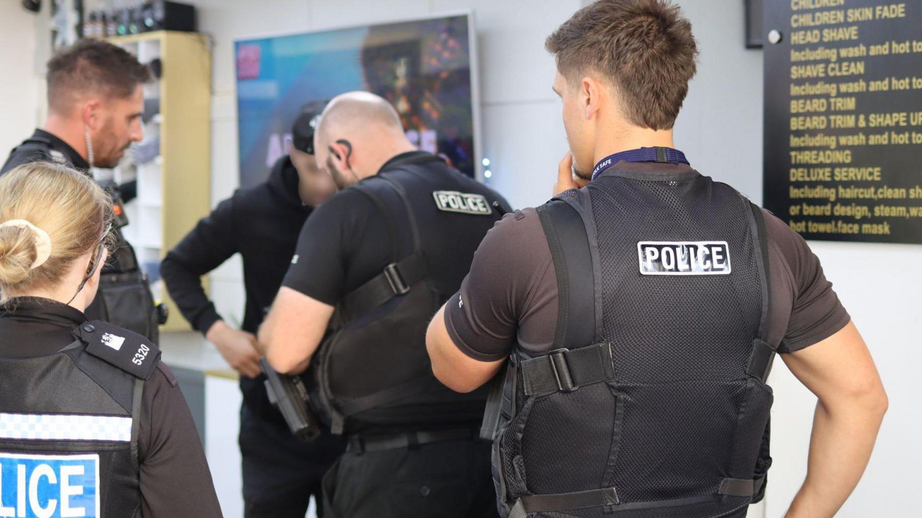 Four police officers are talking to a man who has had his face obscured. Their backs are to the camera.  All are wearing black shirts and stab vests.