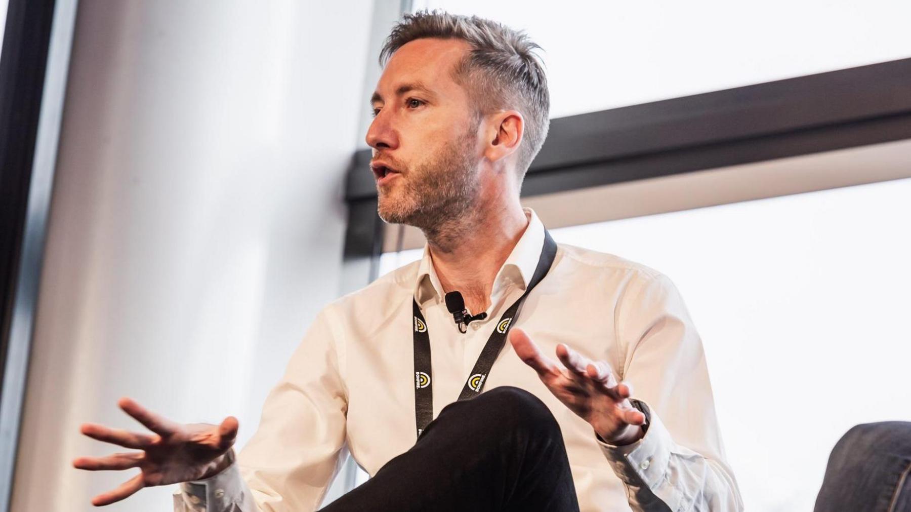 A man wearing a white shirt, black lanyard and microphone attached to his collar is giving a talk at an event which looks like a conference.