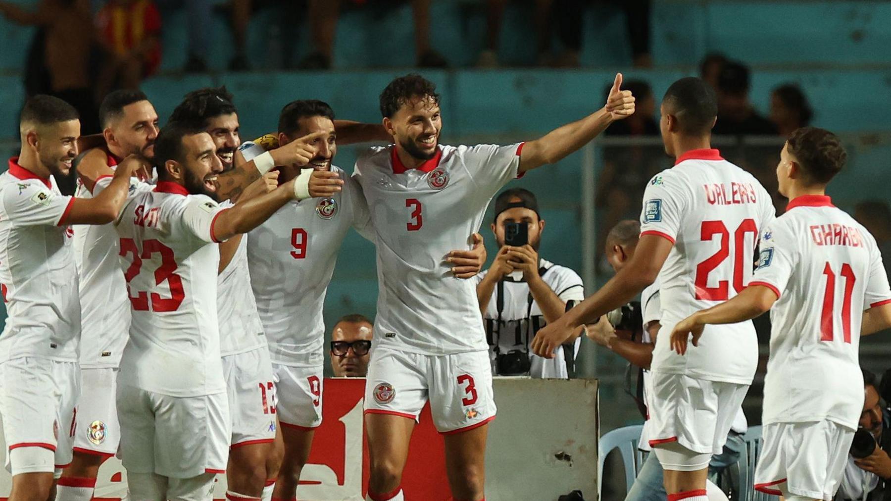 Eight Tunisia players, wearing an all-white strip with red numbers and trim, celebrate a goal with a group huddle as a photographer with a camera on a strap around his neck films on a smartphone behind an advertising hoarding