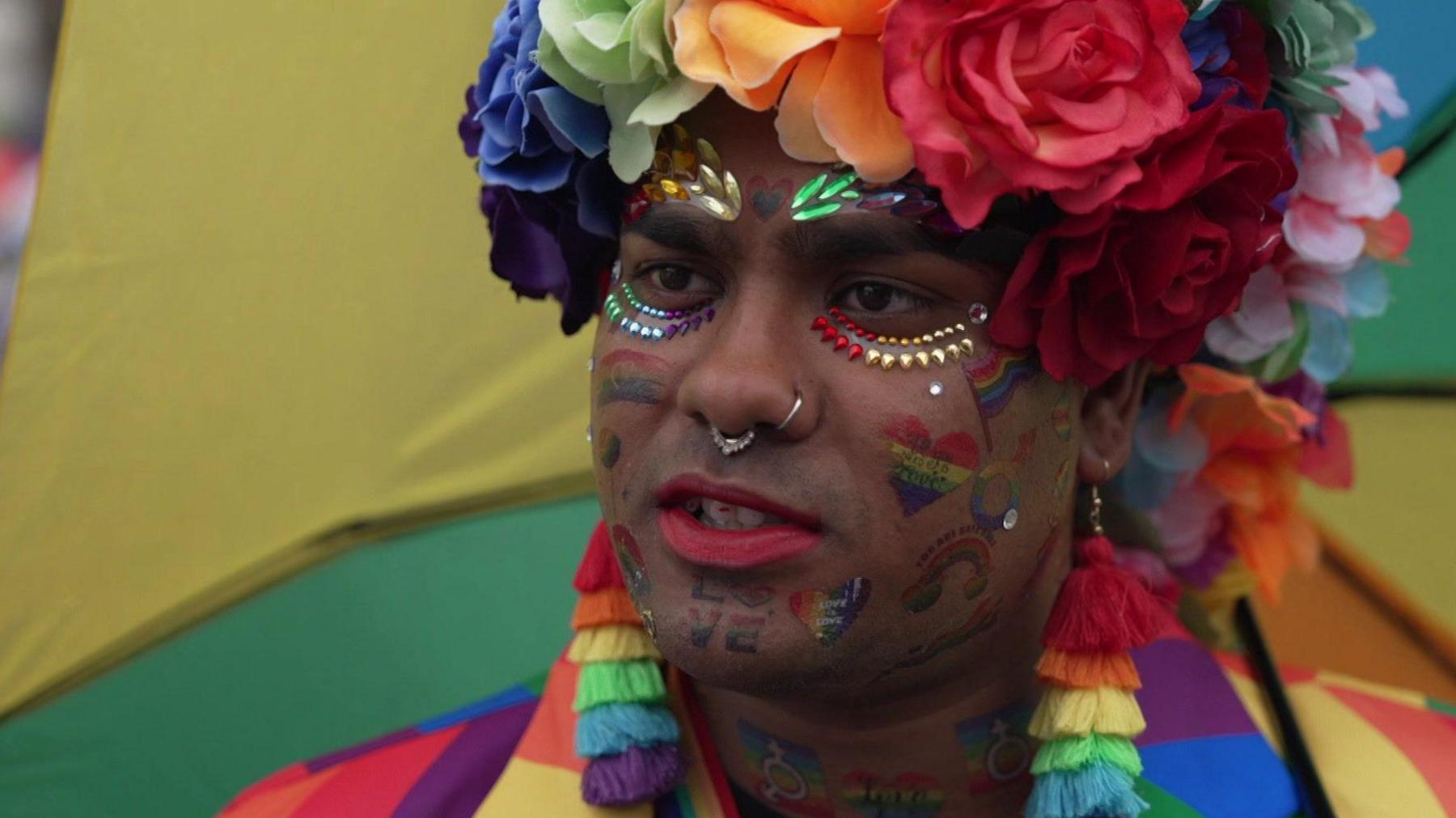 A man wearing multiple brightly coloured flowers in his hair, paintings and piercings on his face and dangly rainbow-coloured earrings.