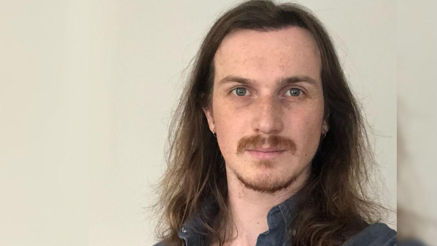 Kieran Moulton wearing a blue collared shirt and standing against a white wall. He has long brown hair and a moustache