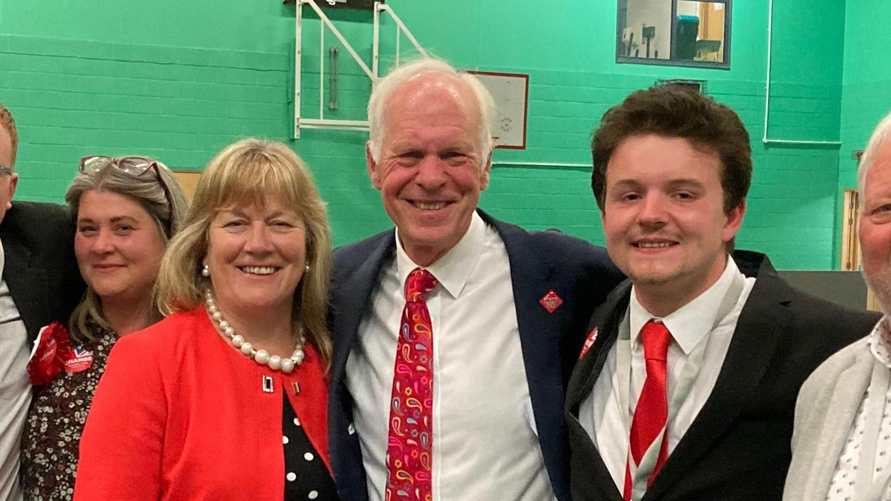 Sir Nic Dakin flanked by supporters on election night, he is smiling. A woman on the left is wearing a red jacket and a chunky necklace. The man on the right is wearing a dark suit and a white shirt with a red tie. 