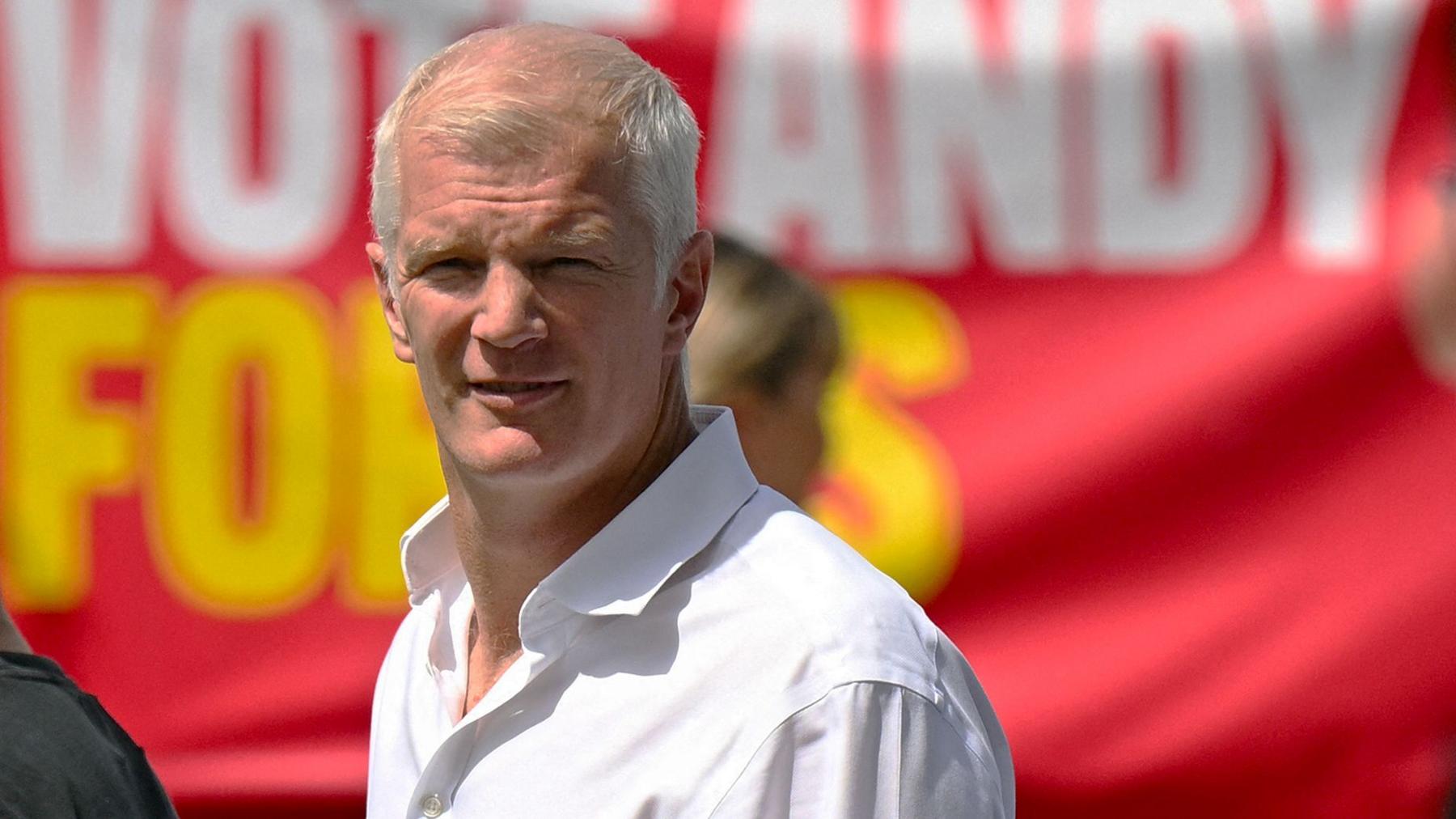 Al Carns outside on a very sunny day in a white shirt against a red banner looking at the camera.