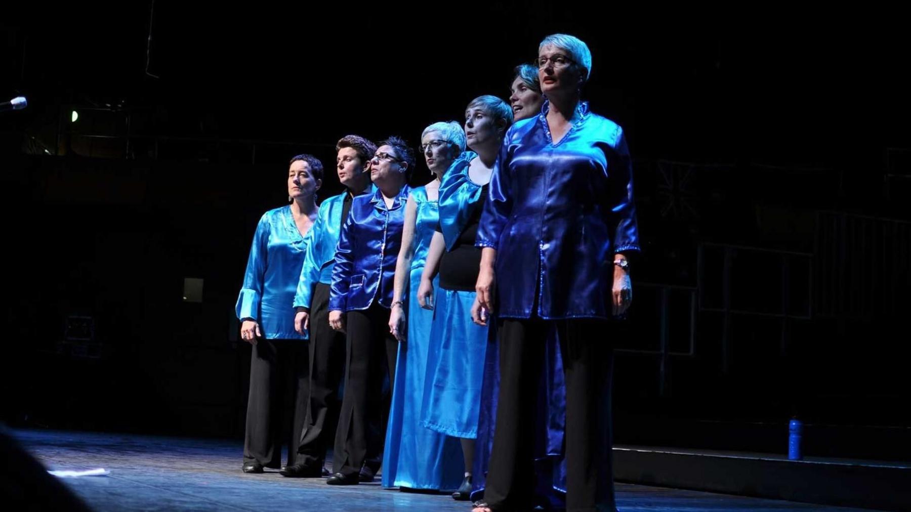 Seven people stand in a line on stage singing in blue outfits