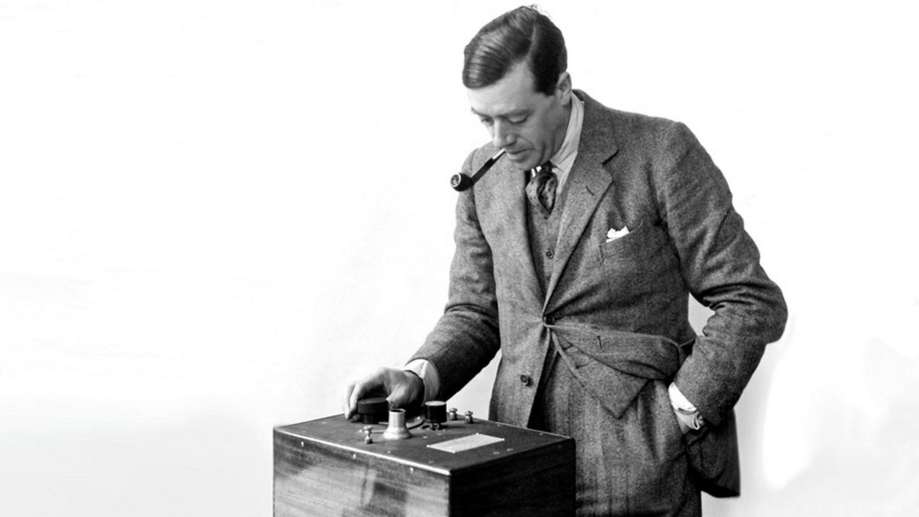 Peter Eckersley with short dark hair smoking a pipe and wearing a grey three-piece suit. His left hand is in his pocket, while the right is touching a switch on a small stand in front of him.