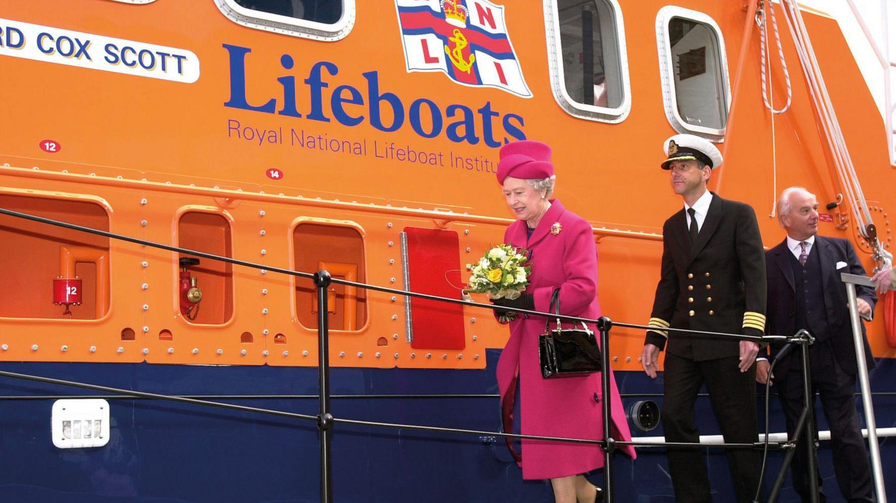 Her Majesty Queen Elizabeth II going aboard Richard Cox Scott accompanied by Falmouth’s harbour master captain Mark Sansom and RNLI chairman Peter Nicholson on 1 May 2002. They are walking along the lifeboat which is orange and blue.