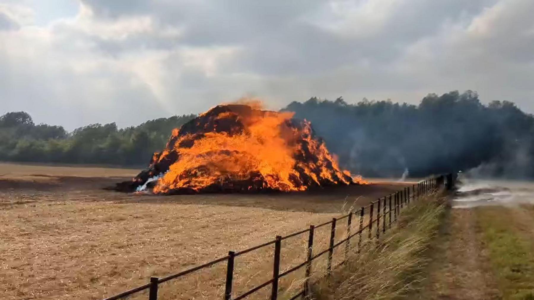 A large pile of hay which is on fire behind a wooden fence with fields all around.