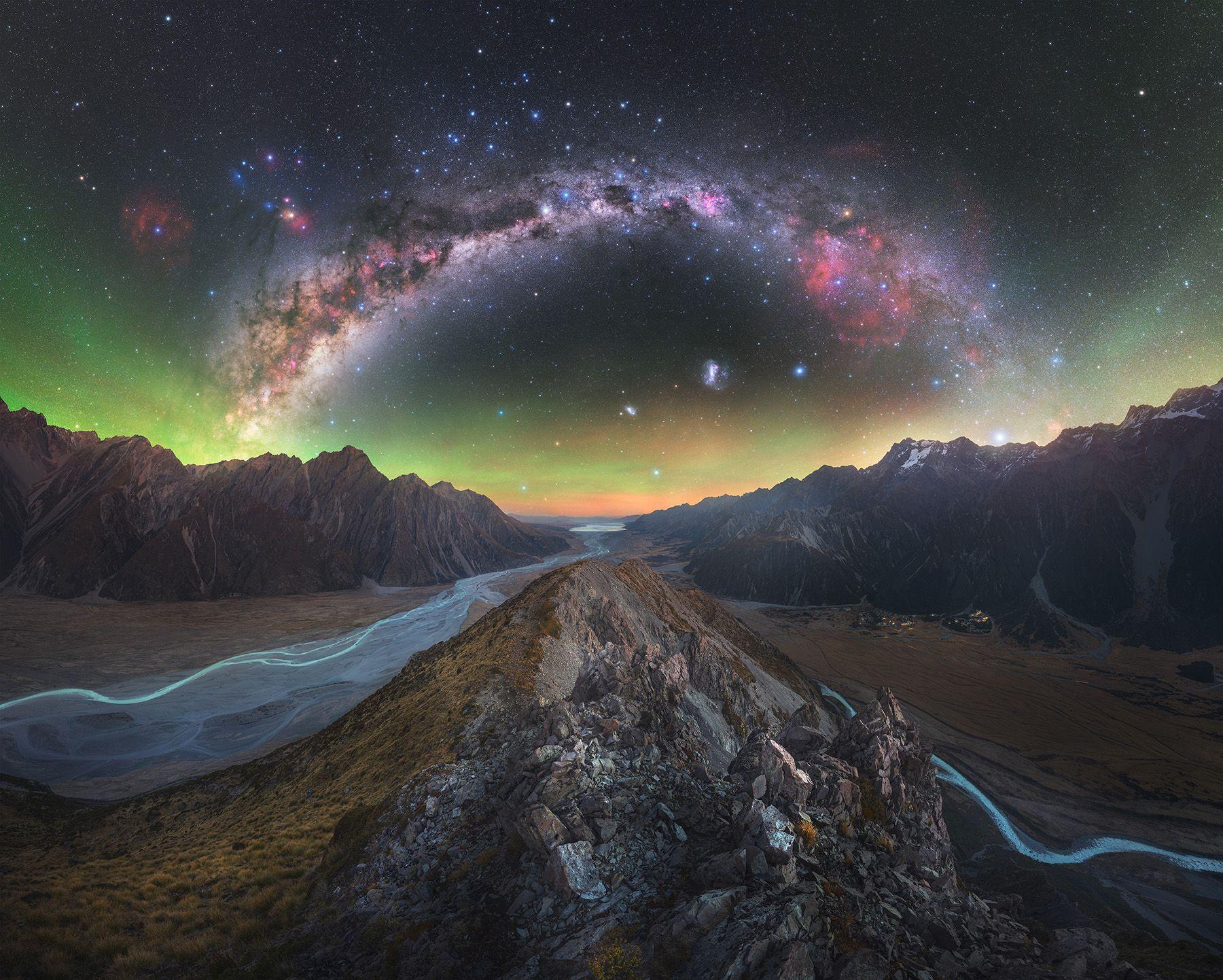 The colourful milkyway and northern lights fill the top half of the image, whilst the bottom half shows a rocky ridge with two very blue rivers running either side.