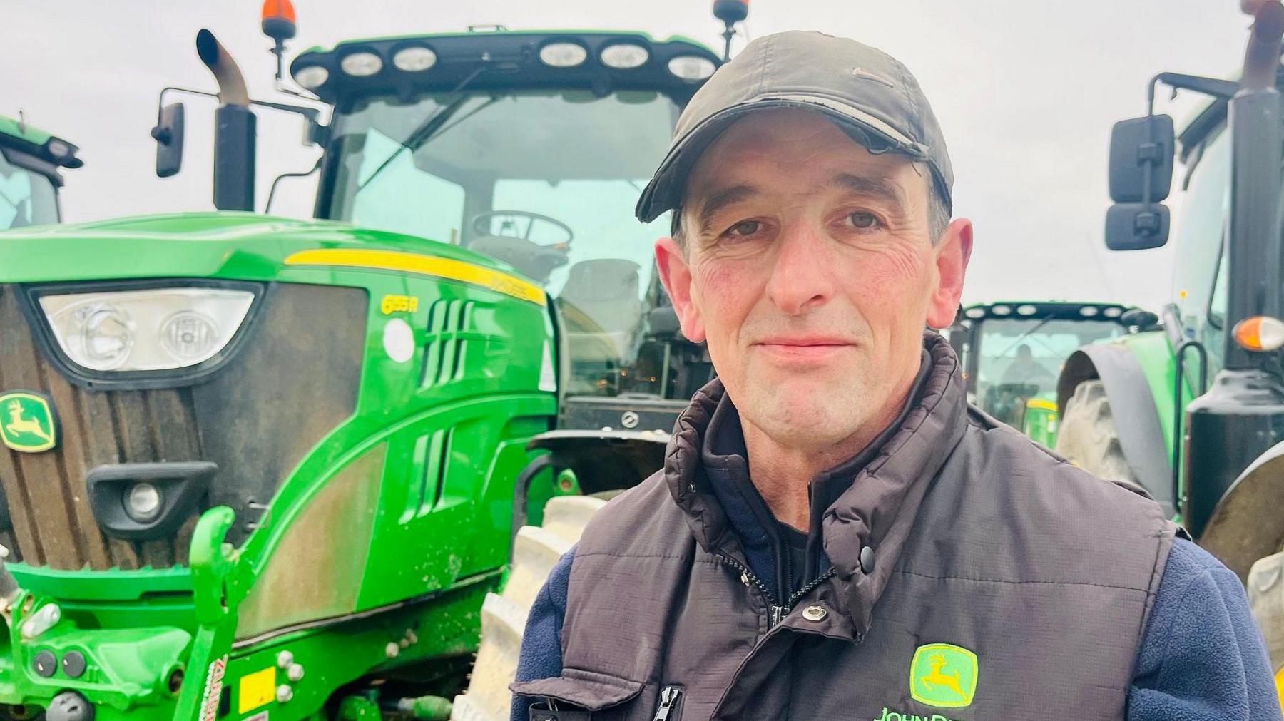 A man stands in front of several green tractors. He is wearing a black gillet, blue fleece and a black baseball cap.