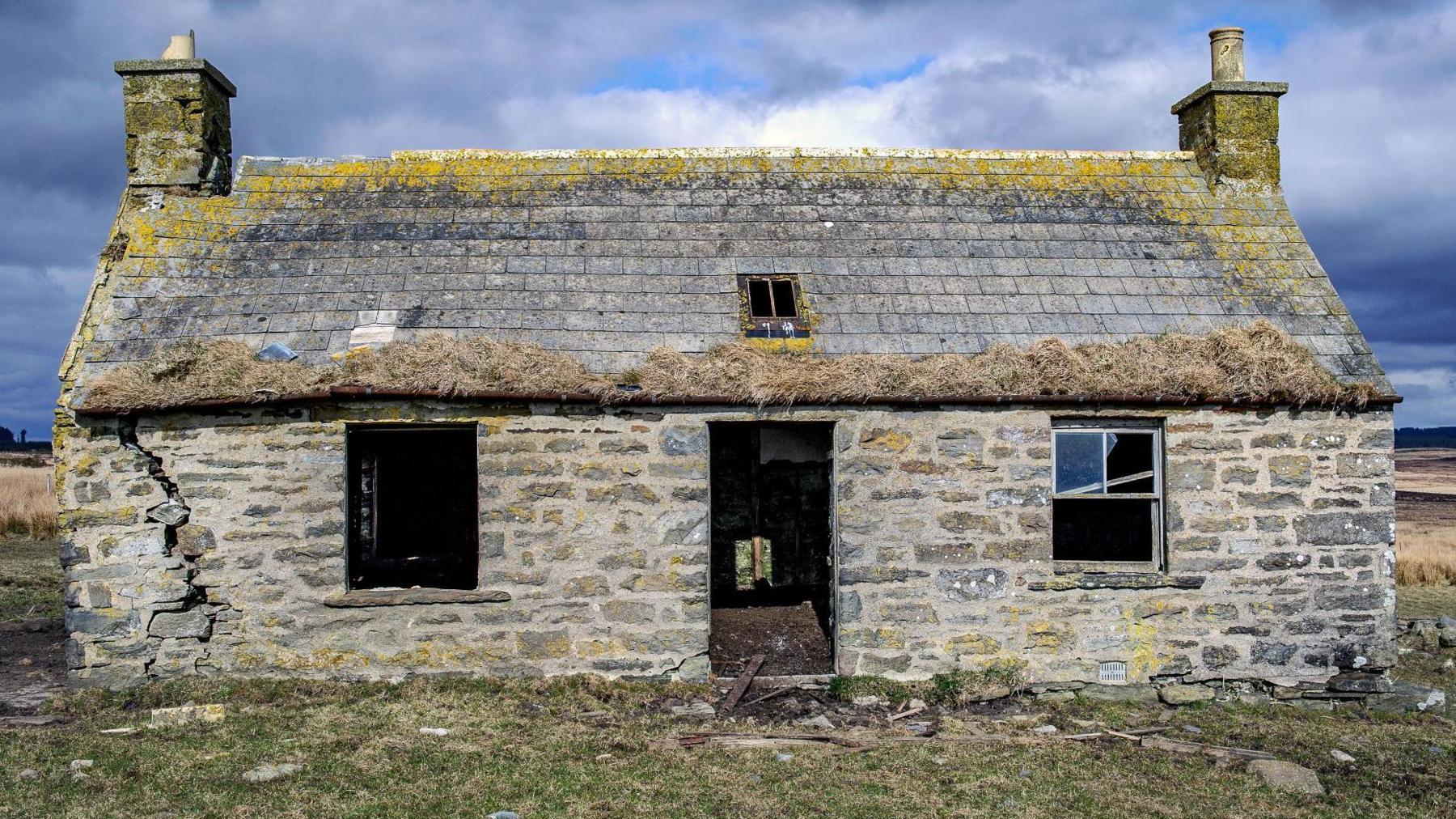 The cottage has a big crack down one exterior wall. There is no front door or glass in the windows. Yellow lichen covers the slate roof.