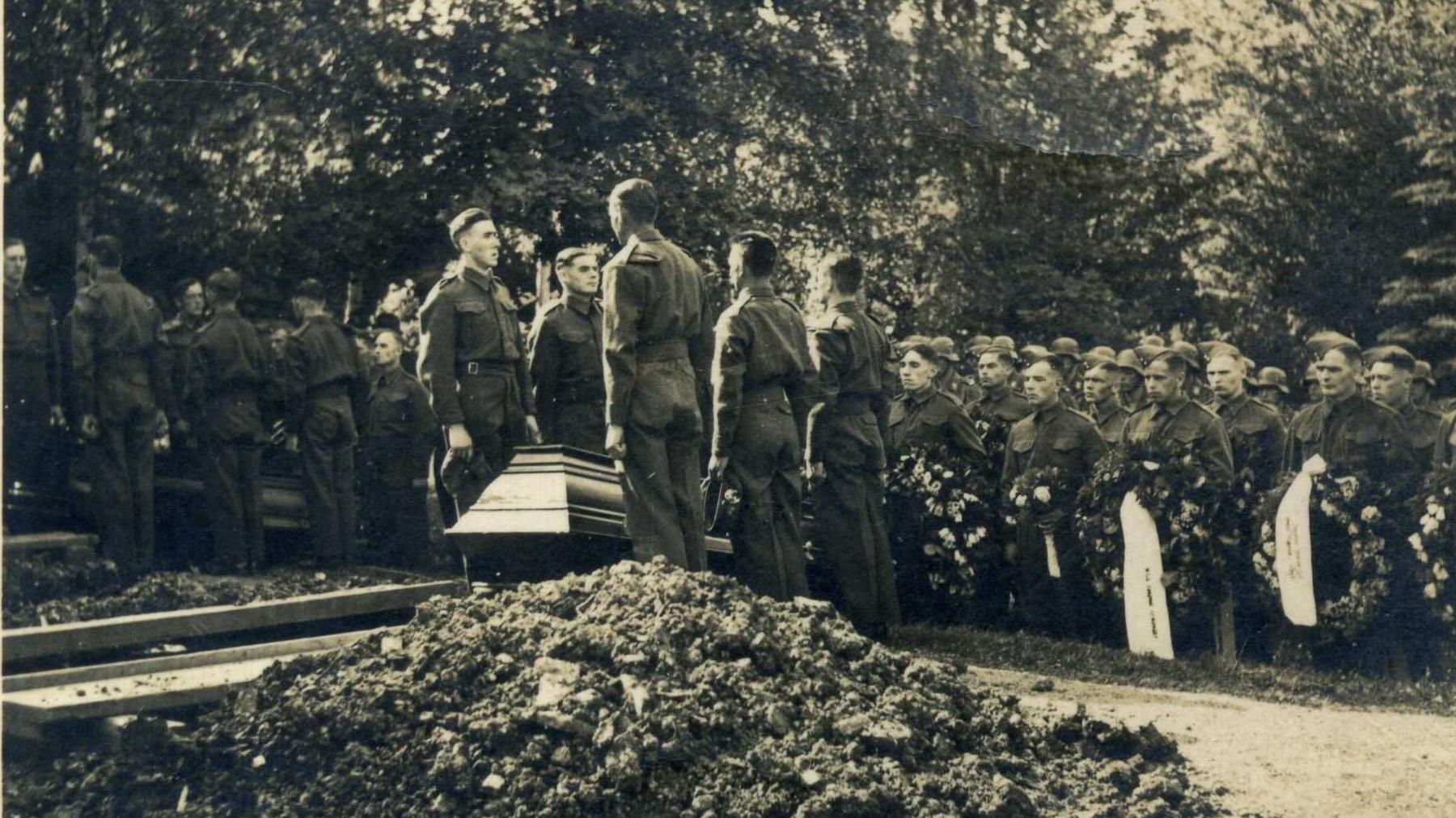 A monochrome photograph from the 1940s which shows a burial ceremony in Berlin. In the foreground there is a pile of earth which has been removed from a freshly dug grave. Five soldiers in uniform can be seen standing to attention by a wooden casket. Further ranks of soldiers holding ceremonial wreaths stand in the background.
