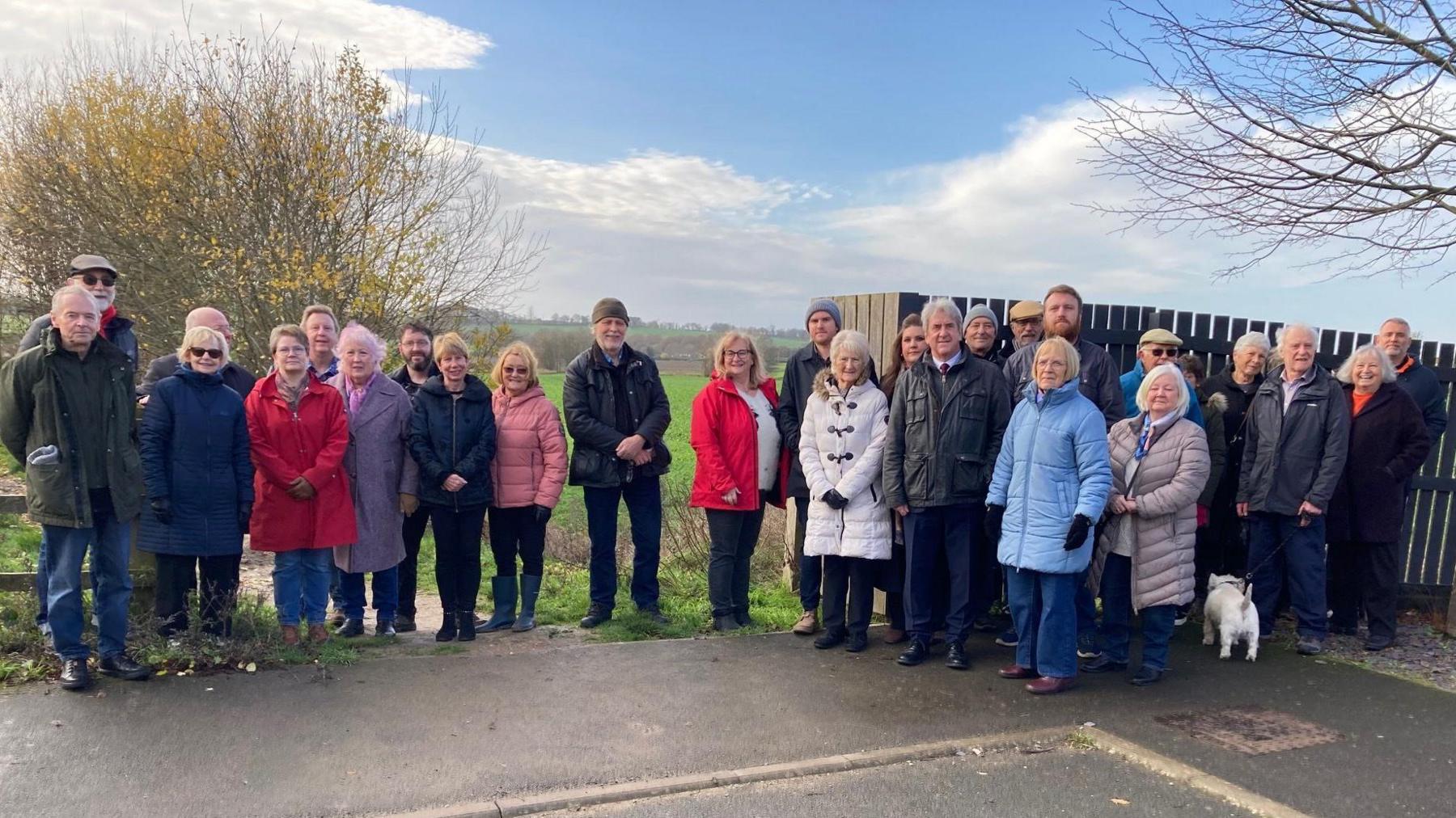A group of people standing together outdoors on a paved area near a wooden fence and a leafless tree. They are dressed in winter coats of various colors, including red, blue, gray, and beige. The background shows a grassy field and trees with autumn foliage under a partly cloudy sky.
