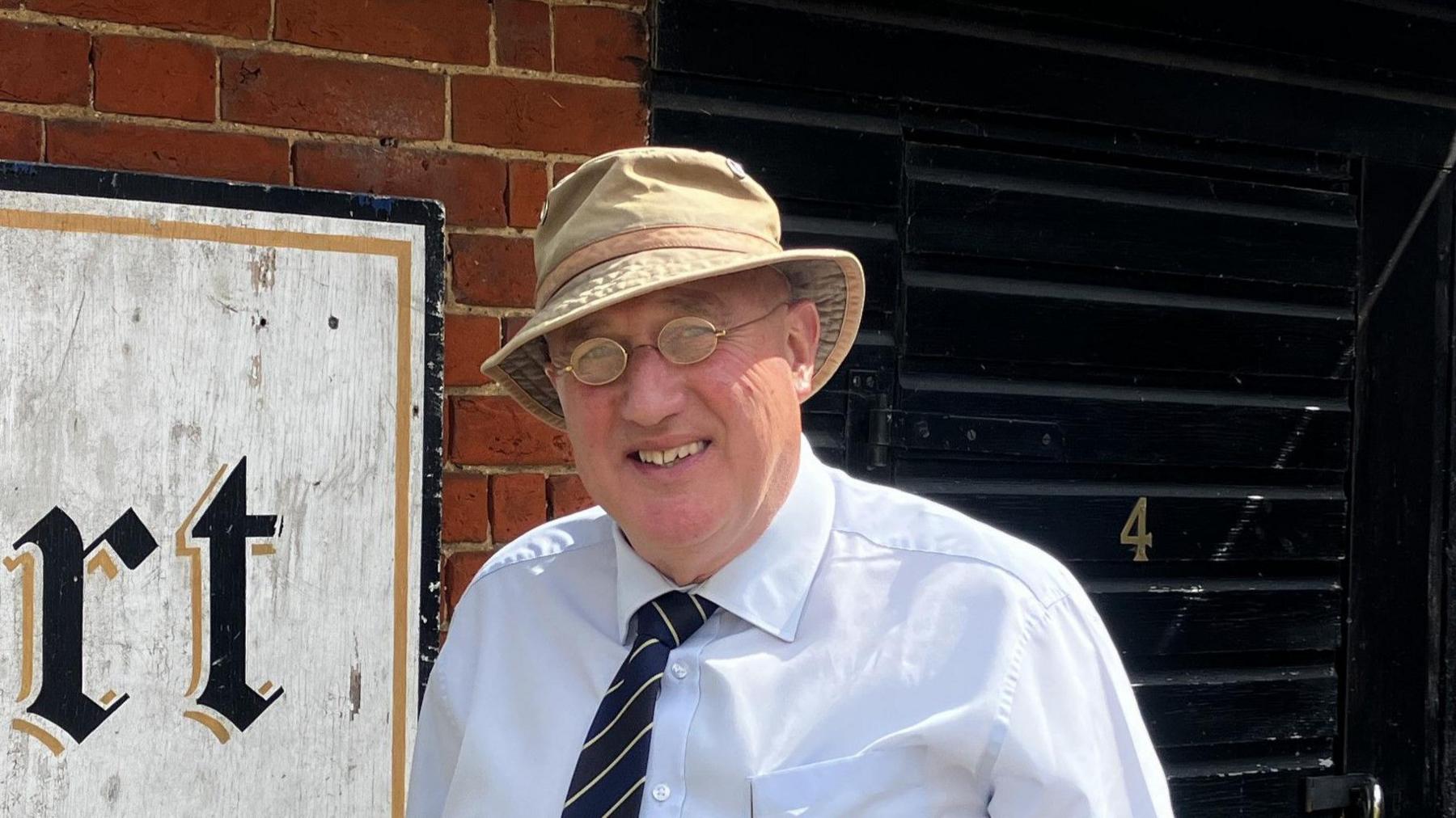 Howard Watts is pictured outside the White Hart pub. He is wearing a cream coloured bucket hat and a light blue shirt with a navy blue and white tie. He has small glasses and is smiling at the camera.