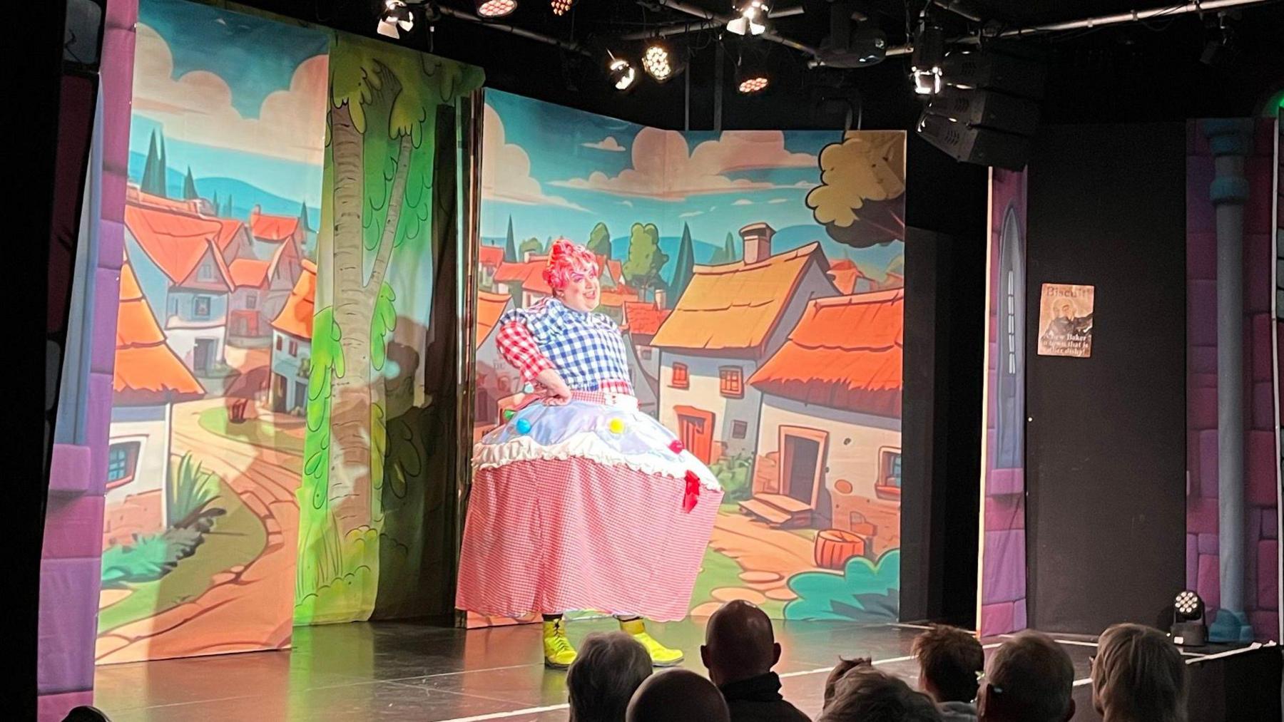 Alex wearing an outfit which looks like a cupcake up on stage, including a blue, white and red chequered blouse, pink wig and a huge pink skirt with a hoop. He is standing on stage in front of scenery that depicts a cartoon village. There are a few audience members looking on in the foreground.