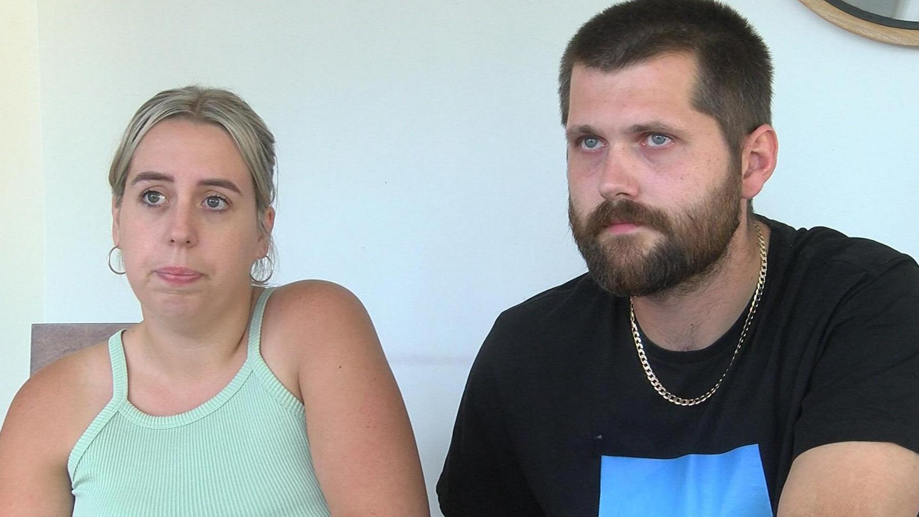 Hollie, wearing light green top and Charlie, bearded, wearing a black t-shirt, sitting in front of a plain light blue wall 