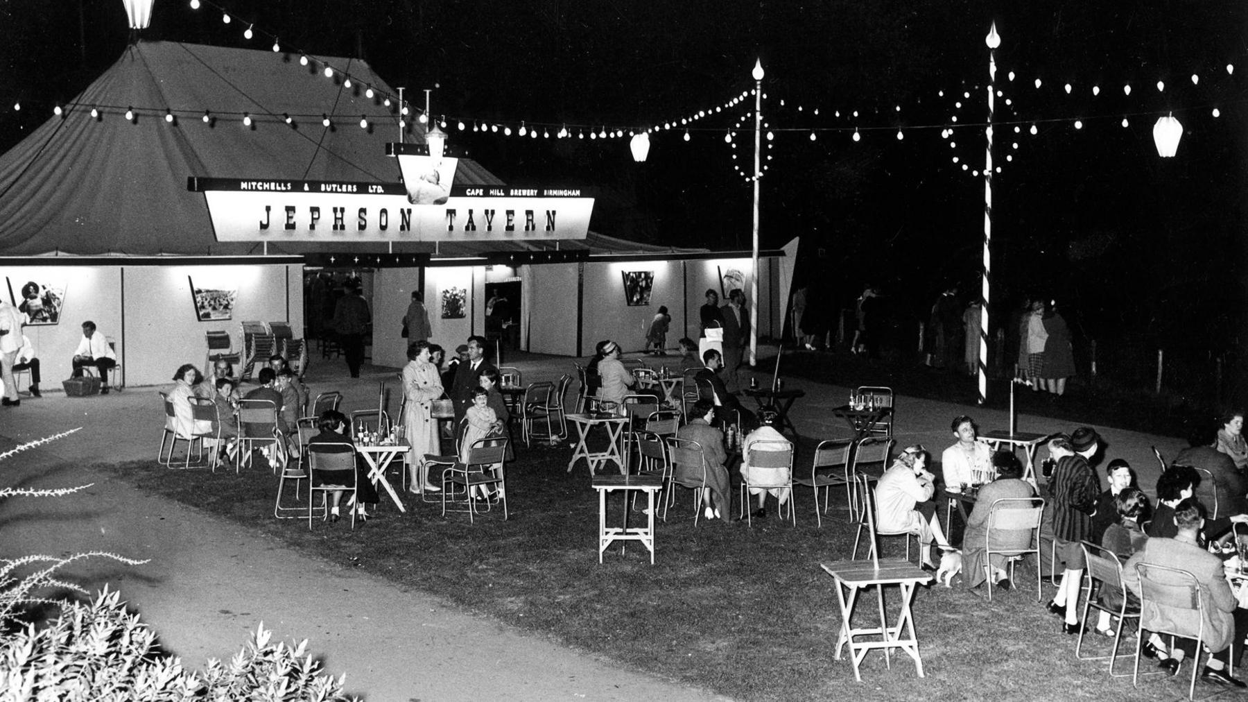 A black and white image showing people sat outside a marquee called Jephson Tavern. They are sat in groups around small tables.