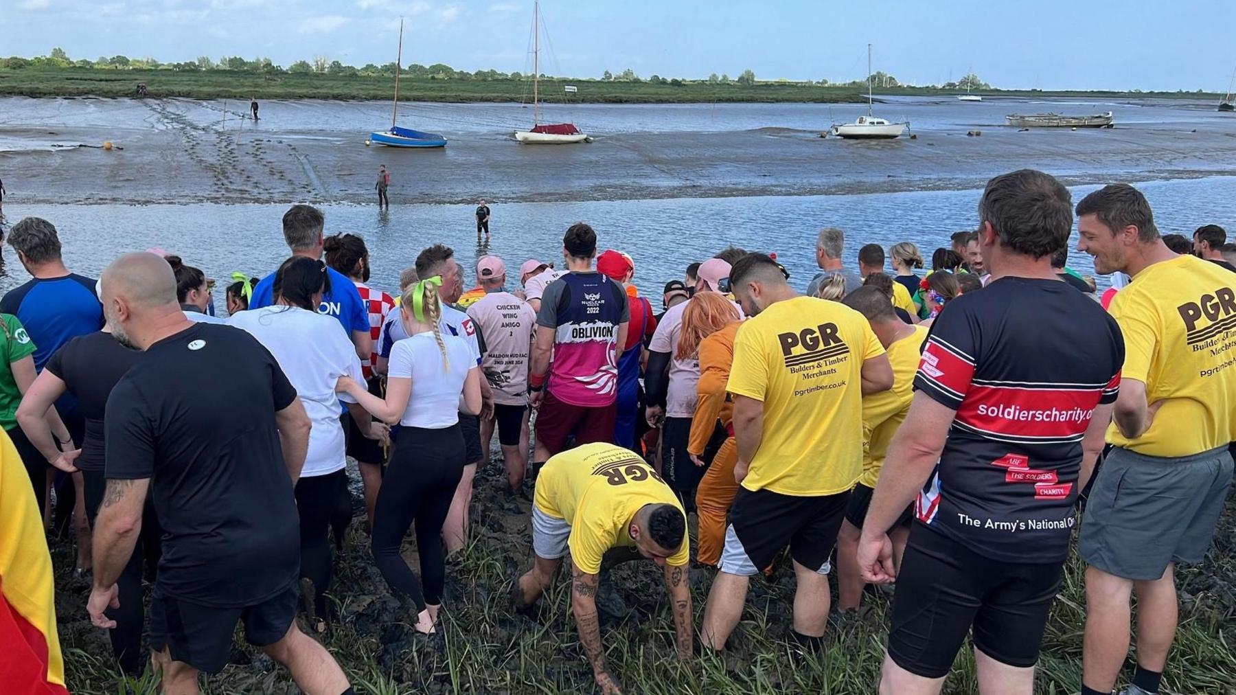 Crowds of competitors lined up on a bank ahead of the race, with the river and mud bank ahead of them, Maldon