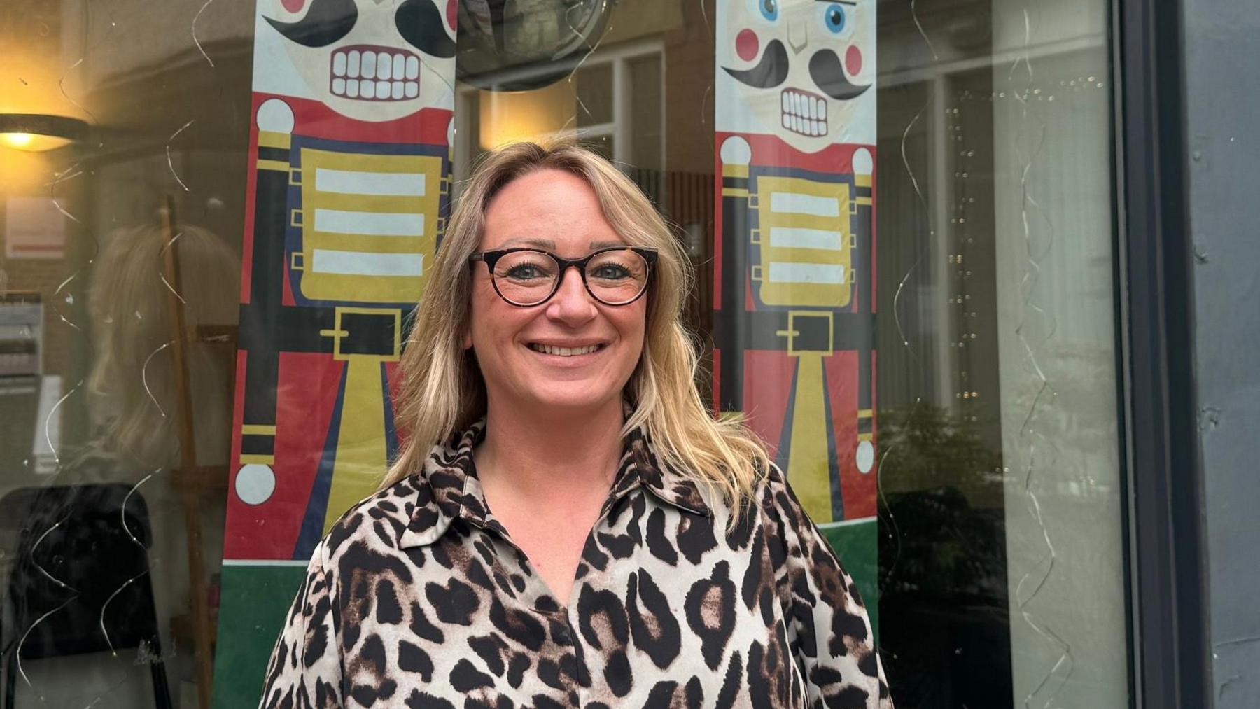 A woman with glasses and blonde hair stands outside a shopfront with nutcrackers in the window.