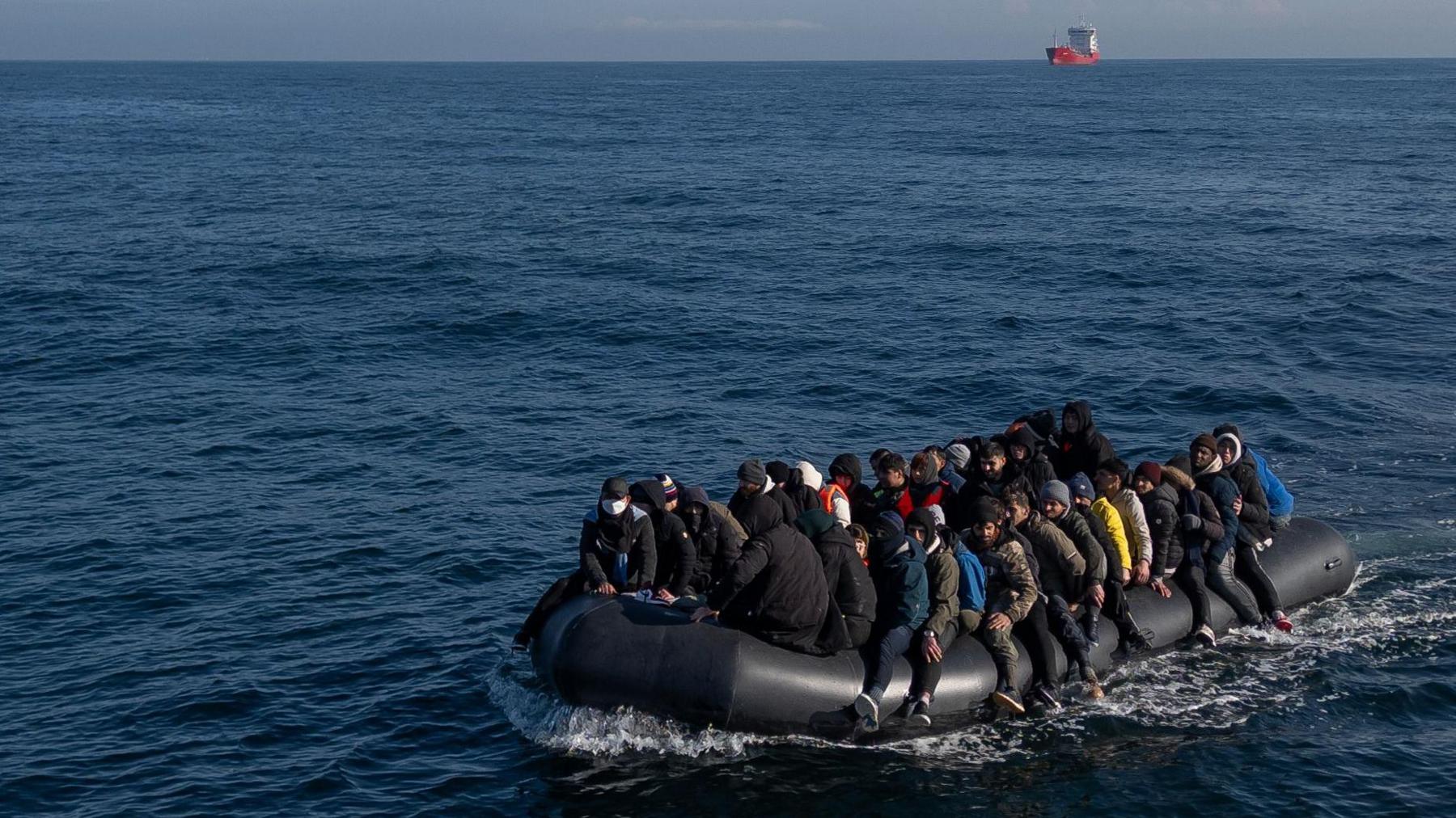 A small boat carrying more than 20 people sails across the Channel, with a larger boat behind it on the horizon in the far distance.