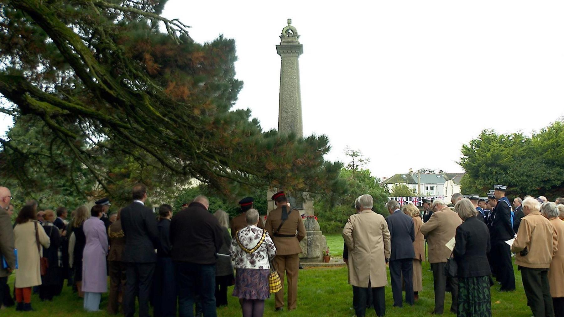 A large group of people in both civilian and military rig stand with their backs to the camera. The are stood outside at Victoria Gardens in Saltash. The group faces a large stone war memorial which has poppies laid at the base. An overhanging tree spans across the left of the image in the foreground. The weather is overcast.