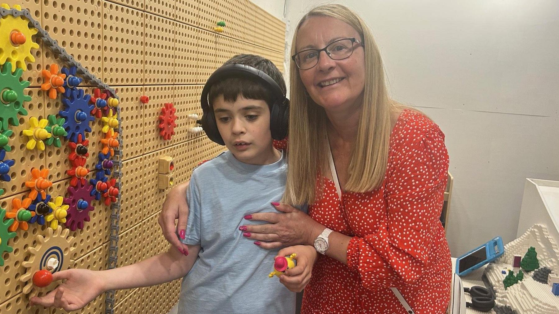 A woman with blonde hair wearing a red spotty dress standing with her son who has headphones on