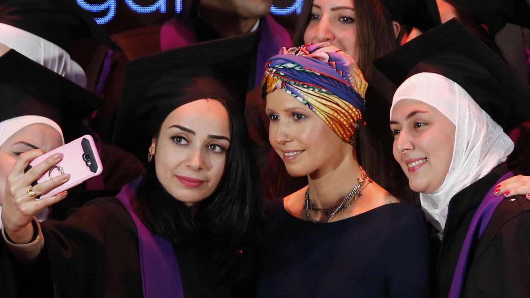 Several women in graduation gowns and caps stand around Asma al-Assad, who wears a colourful head wrap