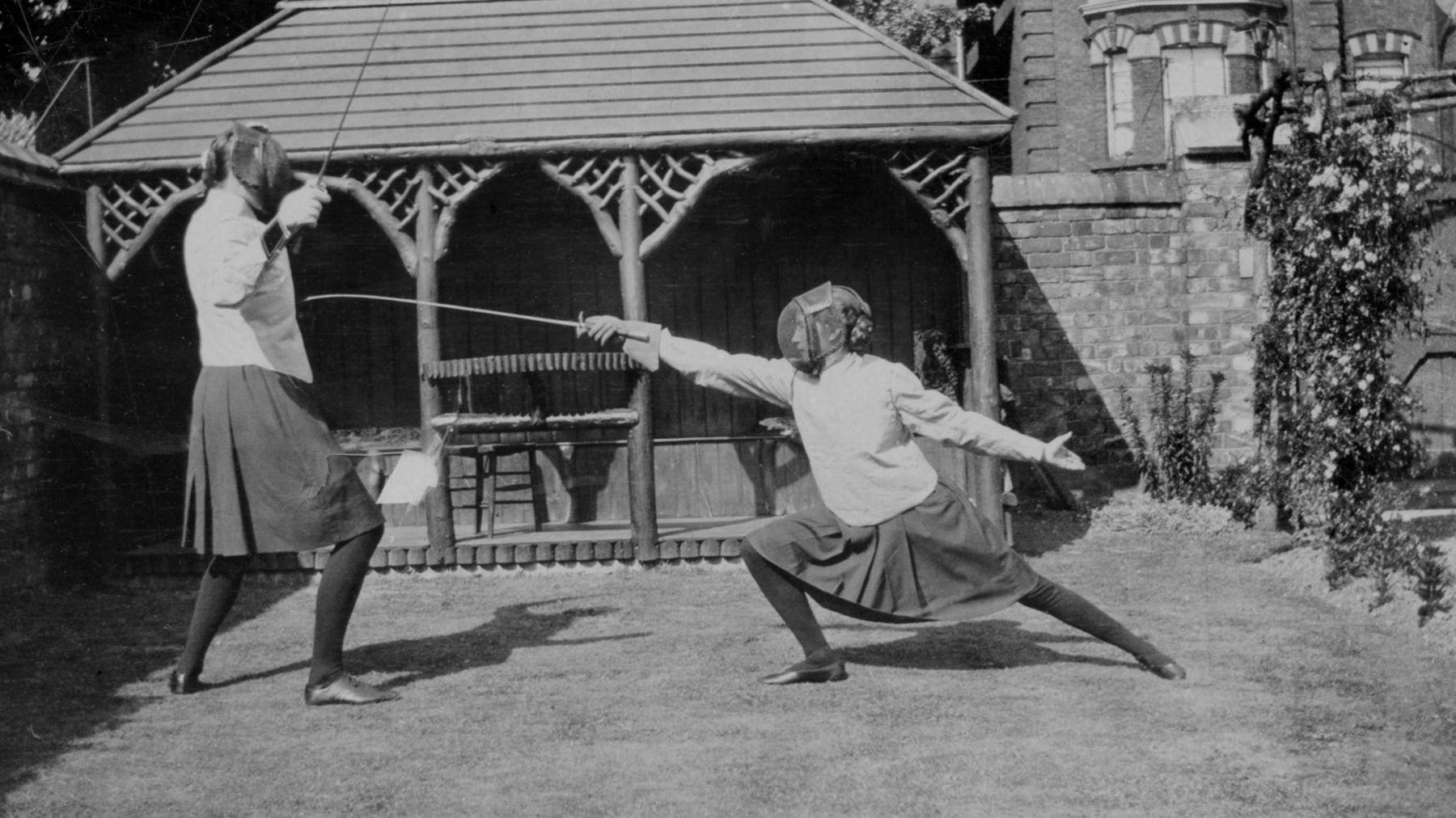 Black and white photo of two women fencing, wearing fencing tops, masks, skirts and tights. 