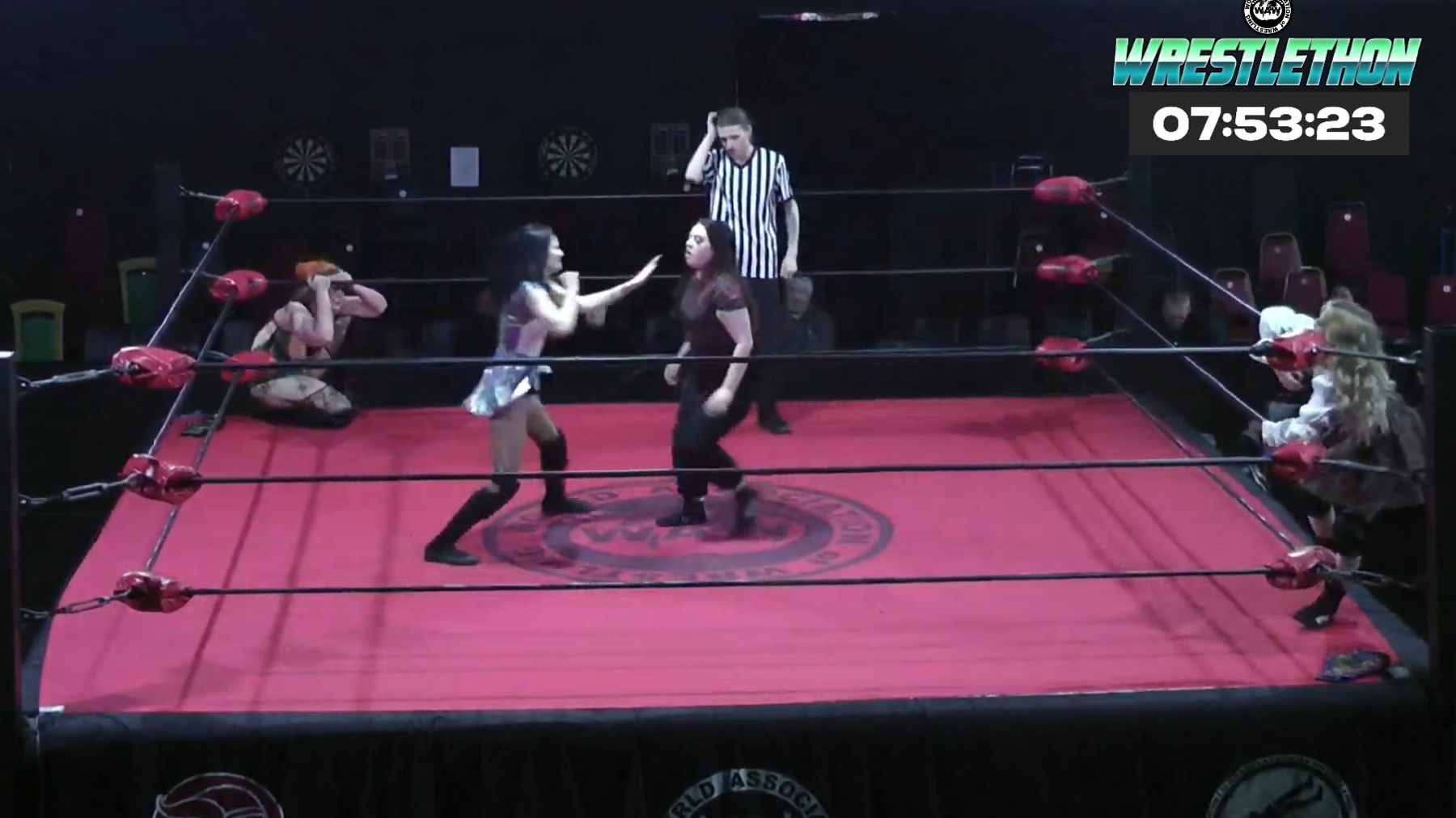 Two female wrestlers in a ring with a red canvas. Two other wrestlers are outside of the ring looking in. A referee in a black-and-white striped t-shirt stands in the ring