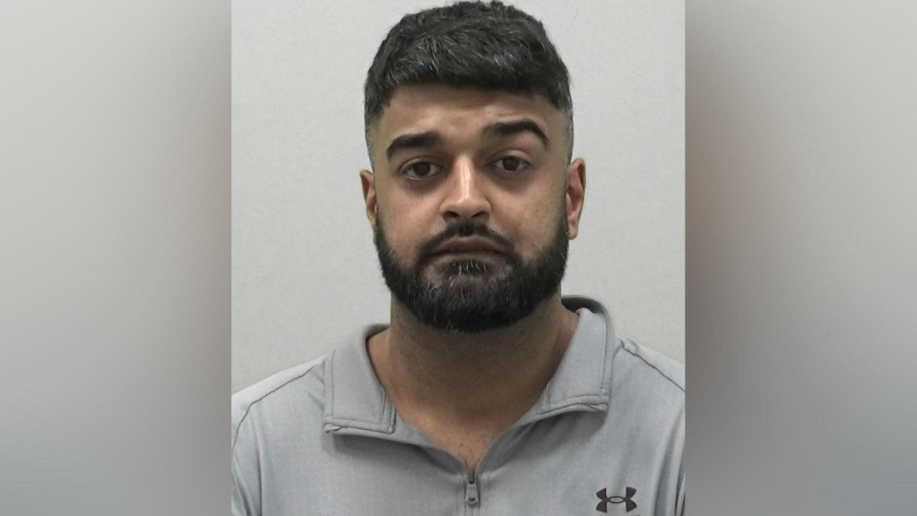 Mugshot of Ali. He has short, neat thick black hair and a black beard and is wearing a grey zip-up polo shirt.