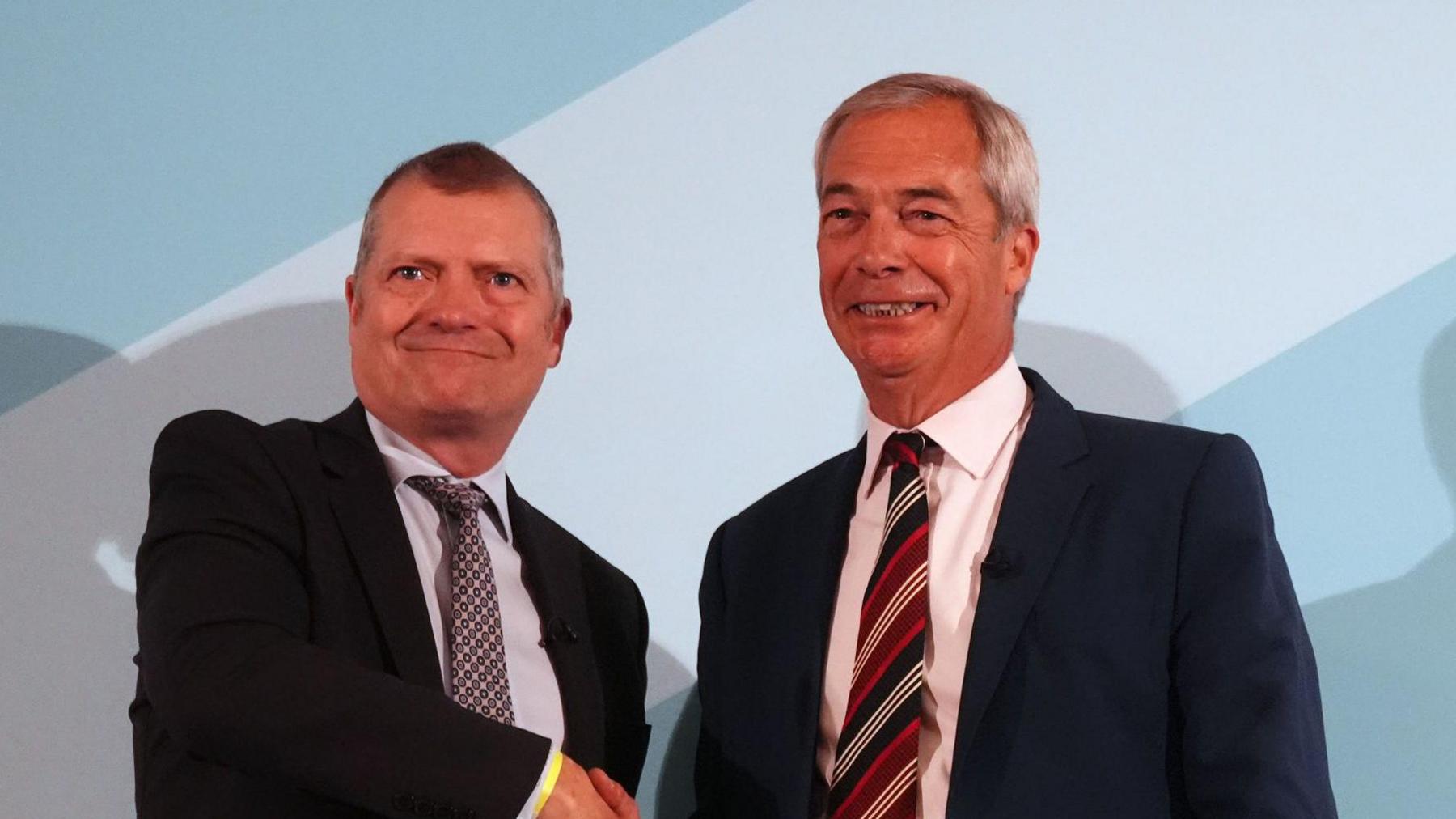 Nigel Farage, with grey hair and a dark blue suit, pink shirt and striped tie is at a podium which says REFORM SCOTLAND. Graham Simpson in grey suit, white shirt and grey tie, shakes his hand