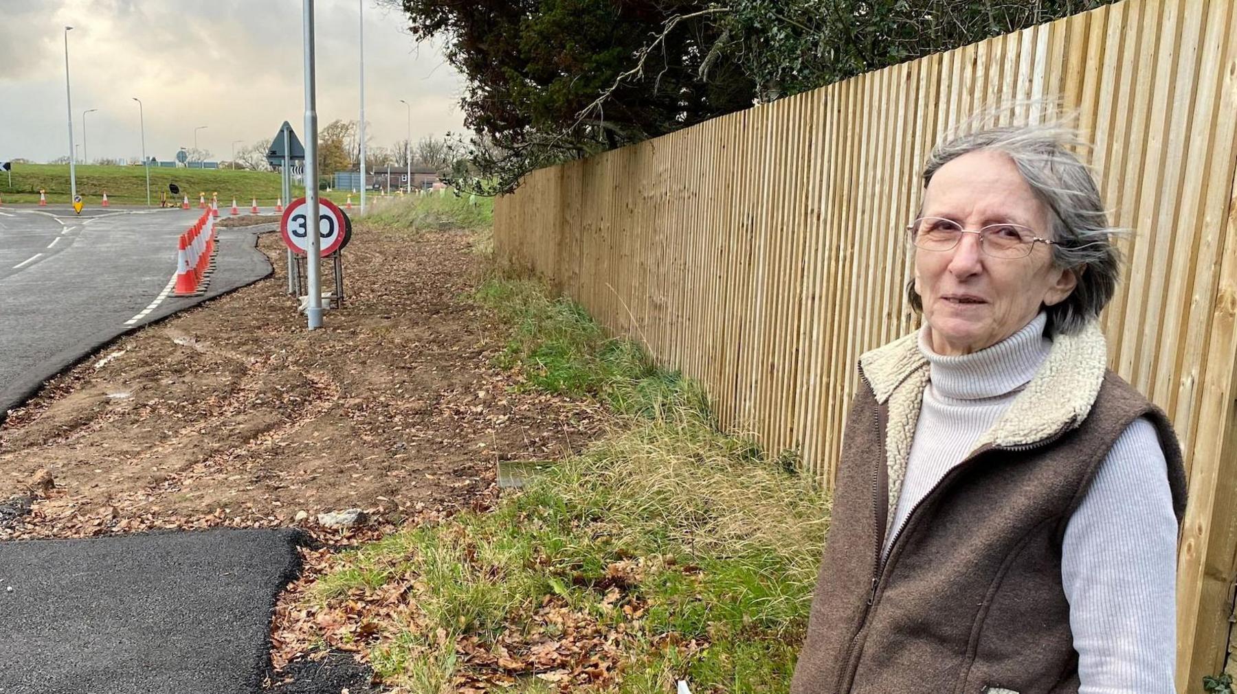 Wendy Pleasent. She has grey hair in a bob and is wearing glasses, a polo neck jumper and sheepskin-type gilet. She is standing by the road which leads to a new roundabout where the statue is due to be placed.