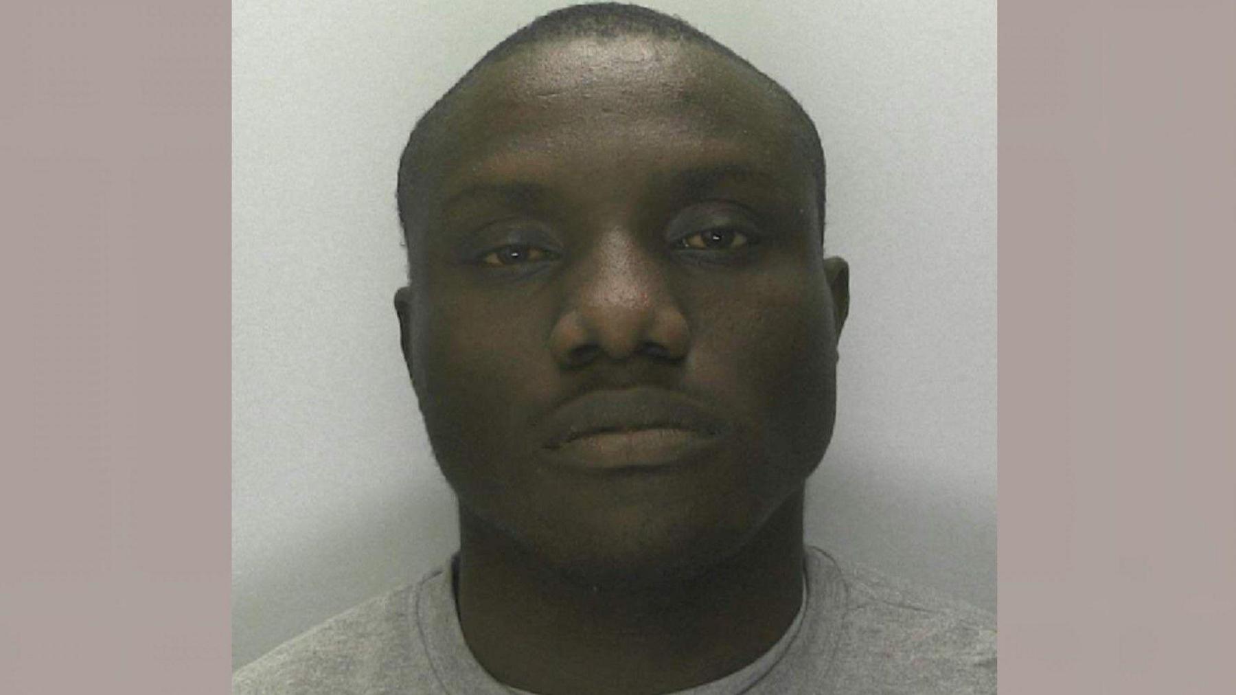 A mug shot of Segin Ogundipe. He is looking into the camera with a blank face and is wearing a light grey sweatshirt