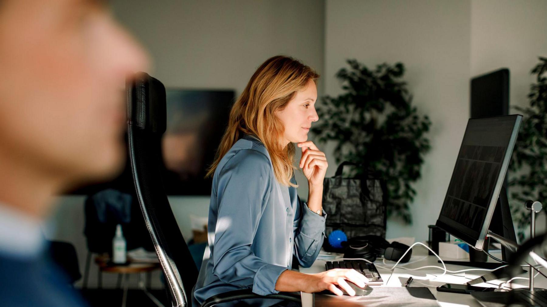 Side view of woman with blonde hair, wearing a blue blouse. Her hand is on her chin while using desktop PC in office 