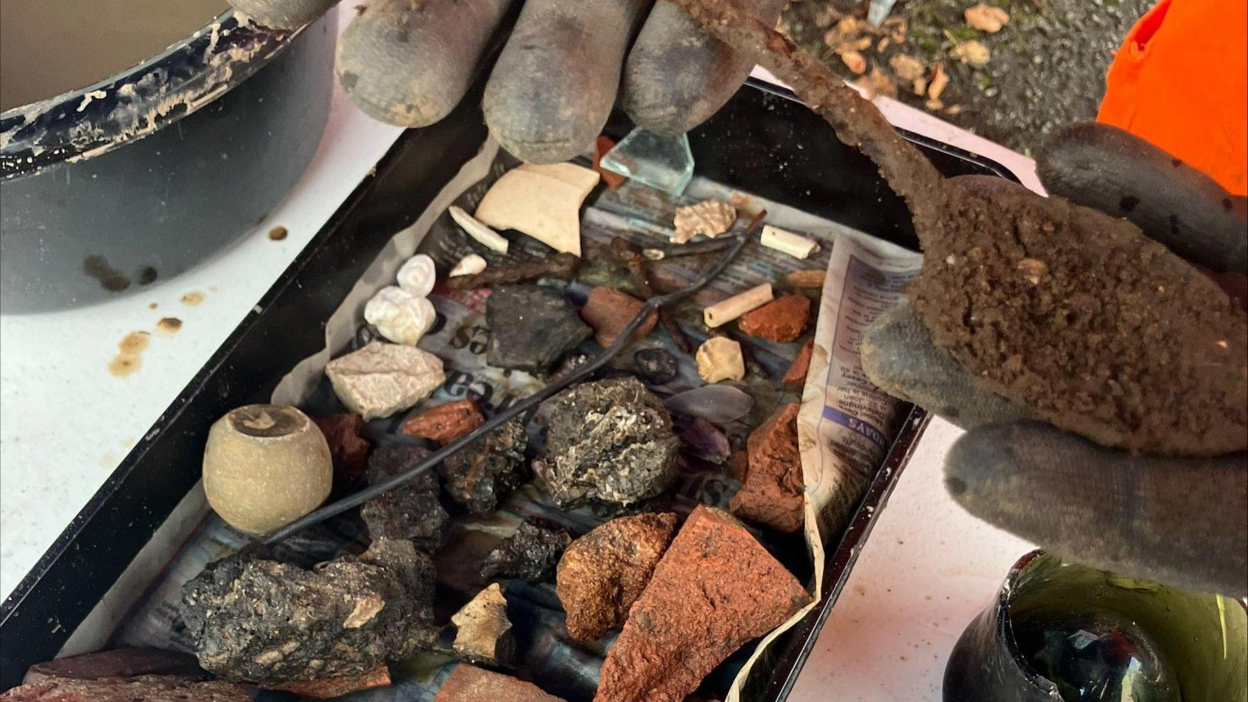 A spoon covered in soil is being held up to the camera by a pair of hands with gloves on. Underneath is a tray with pieces of glass, stone and brick that have been unearthed as part of an archaeological dig.