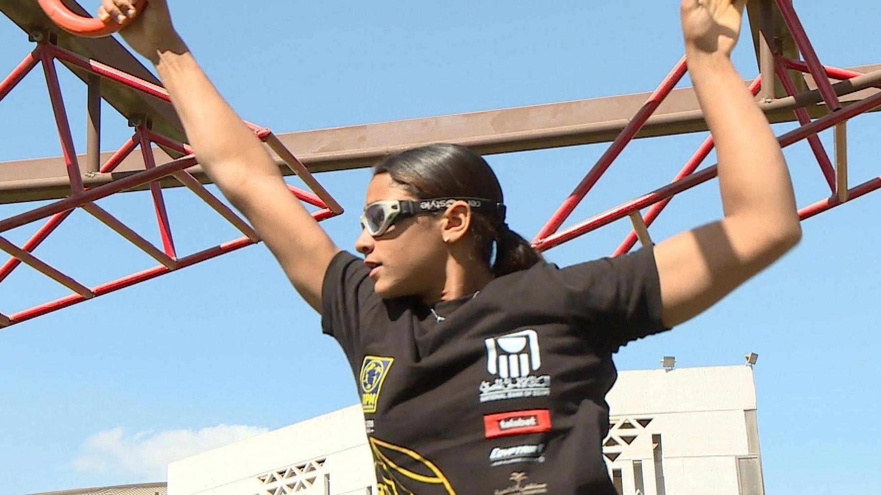 Farida Khalil, visible from waist up and wearing grey and black sports sunglasses and a black T-shirt with yellow detail and sponsor logos on it, is seen midway through an obstacle course. She has her right hand on a red hoop in front of her, and her left hand is letting go of a handhold behind her. The top of a white building can be seen in the distance behind her