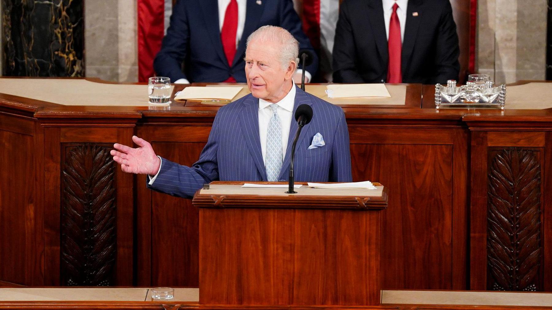 King Charles wears a blue striped suit with a while shirt and light blue tie. He is standing at a podium delivering a speech to Congress at the US House Chamber in Washington. He is gesturing with one hand out to his side