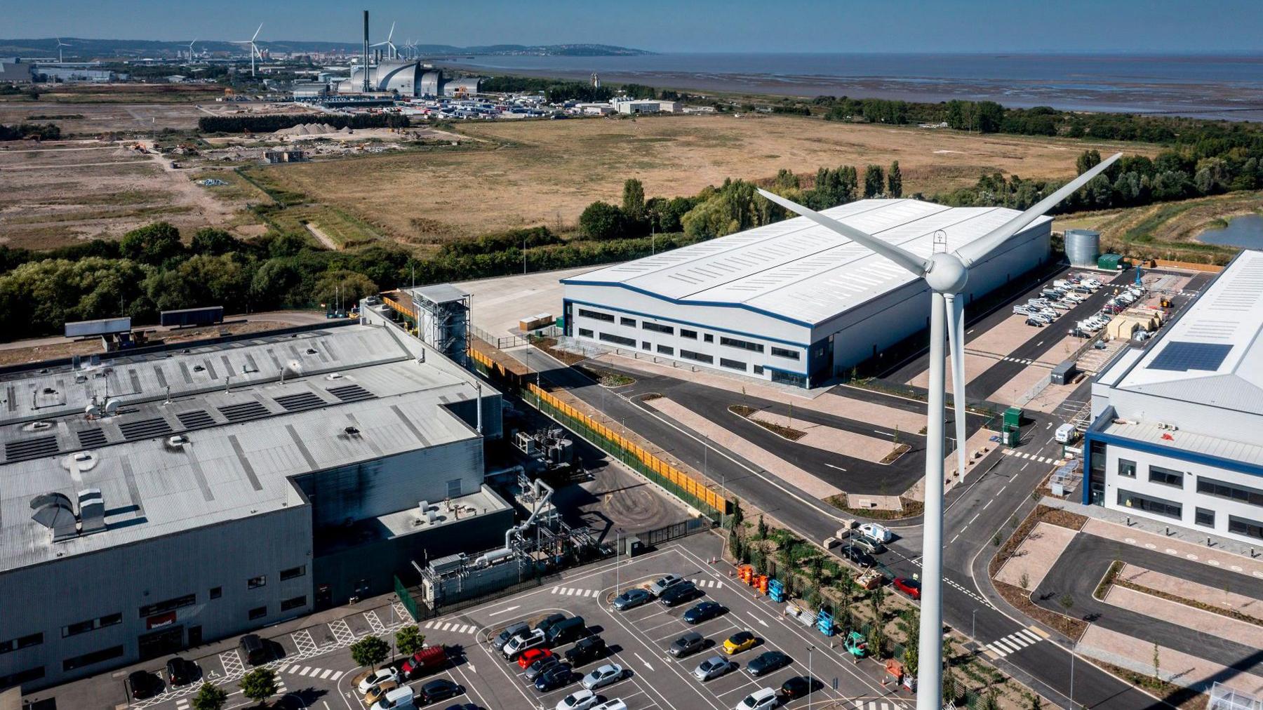 An aerial shot of the new Evri warehouse in Avonmouth. The building is enormous, the scale shown by a wind turbine in the foreground. In the distance other industrial units can be seen along the North Somerset Coast, with the Severn Estuary also visible.